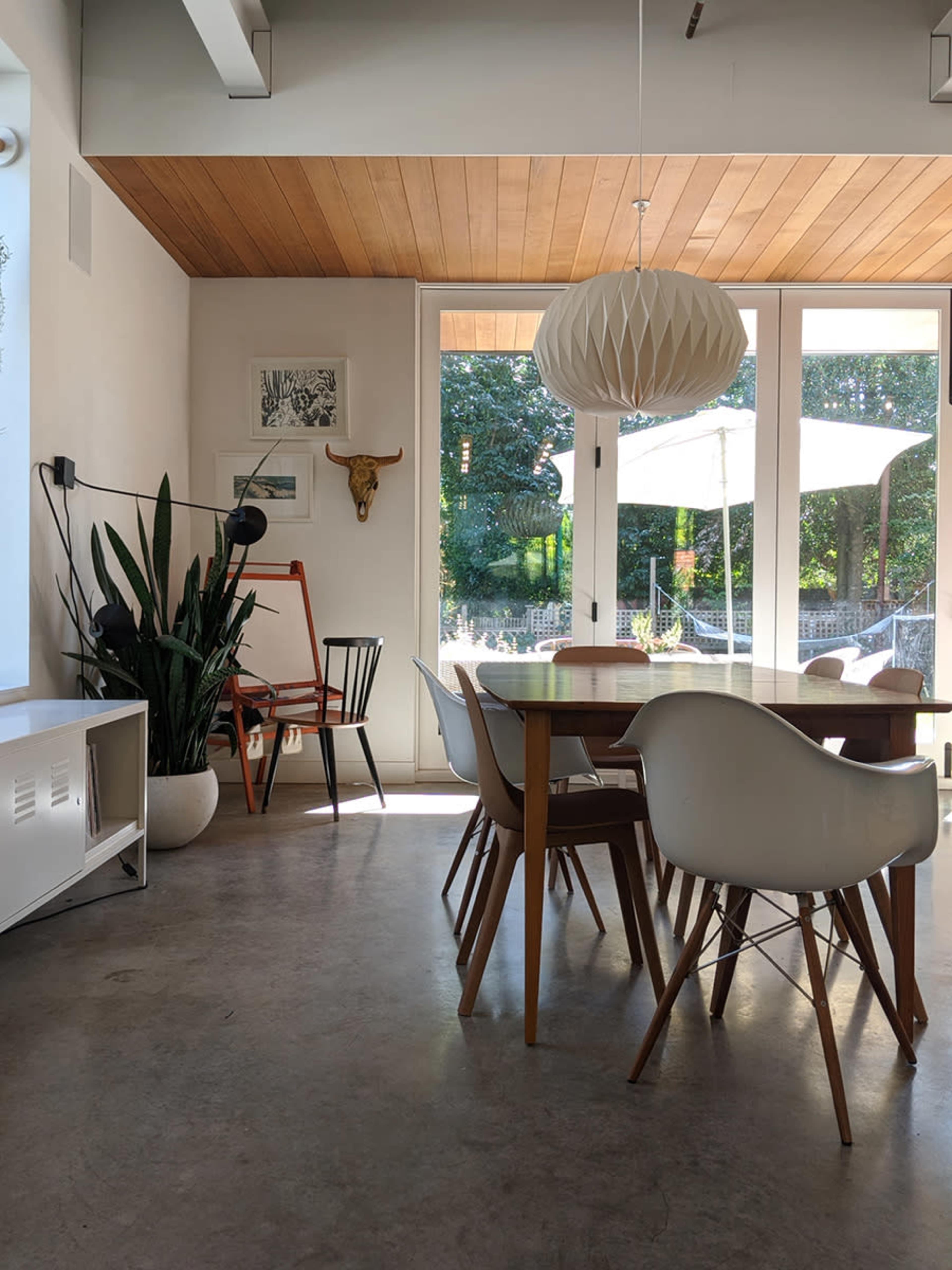 The image shows a modern dining area with a wooden ceiling, a large round light fixture, a dining table surrounded by various chairs, and large windows overlooking a garden.