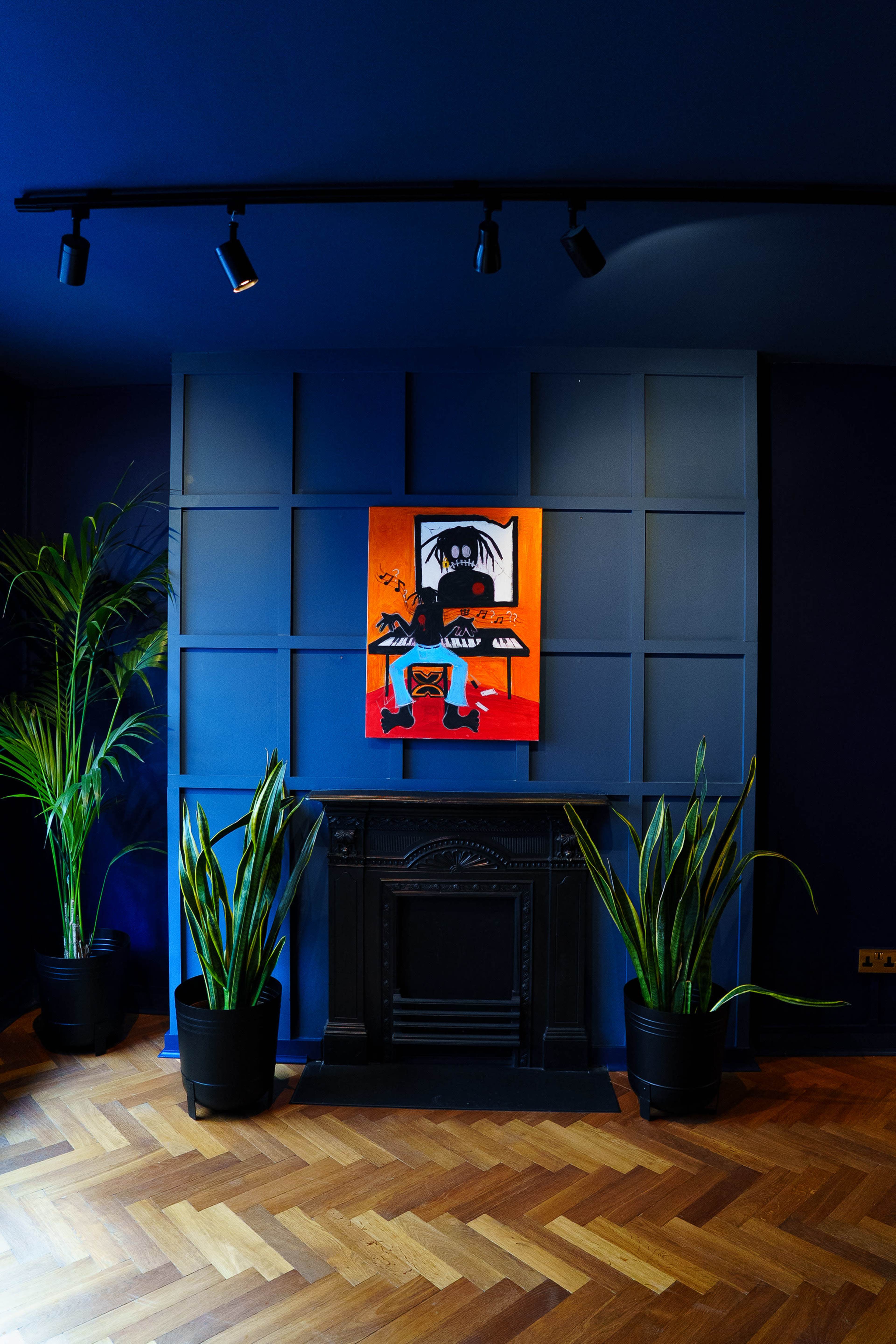 The image shows a modern room with a dark blue wall featuring a colorful painting centered above a decorative fireplace, flanked by tall green plants in black pots.
