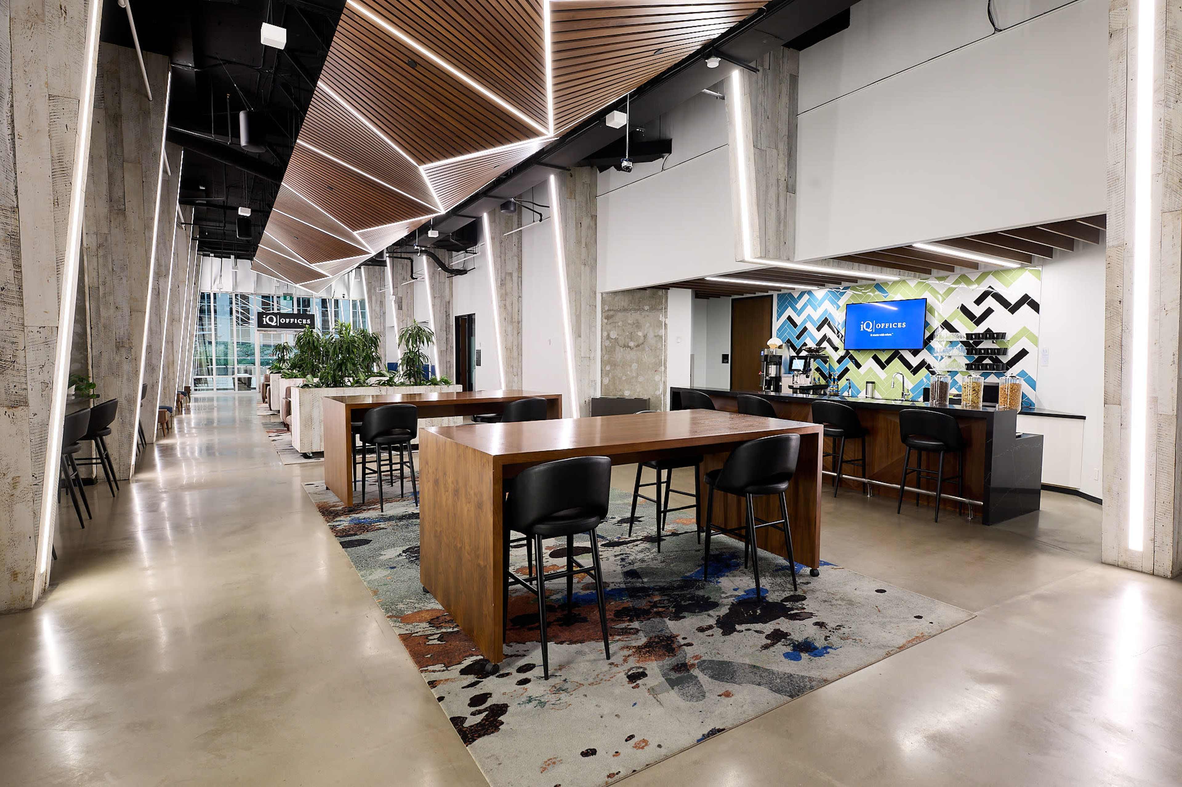 The image shows a modern co-working space featuring large wooden tables, black chairs, and a coffee station, with walls adorned in geometric patterns and greenery throughout the area.