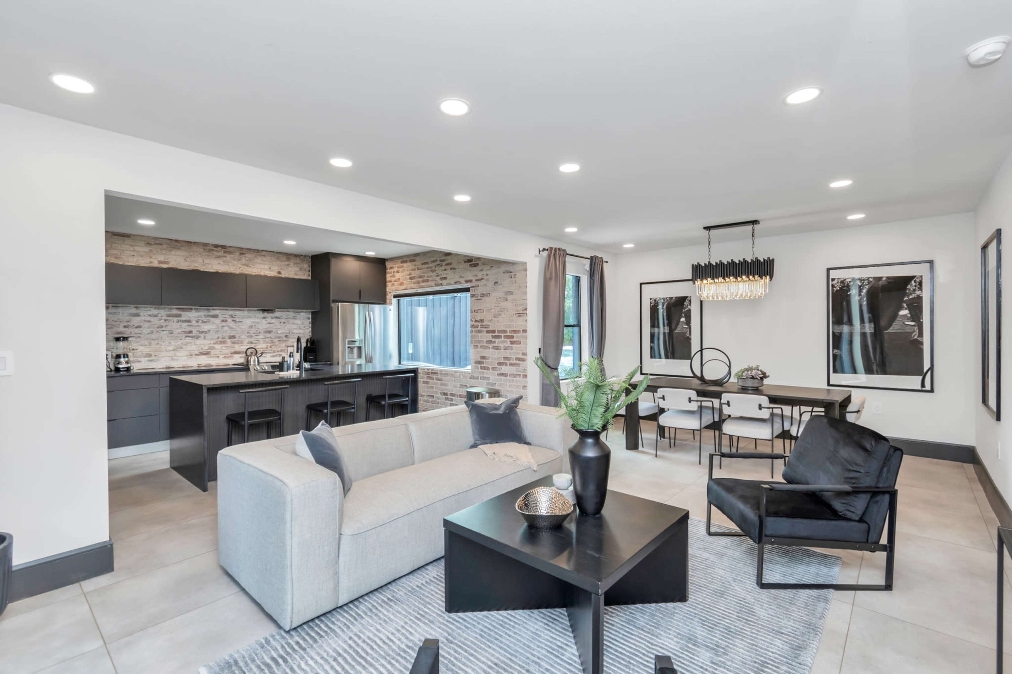 A modern, open-concept living area features a beige sofa, black furniture, a dining table, and a kitchen with exposed brick accents.