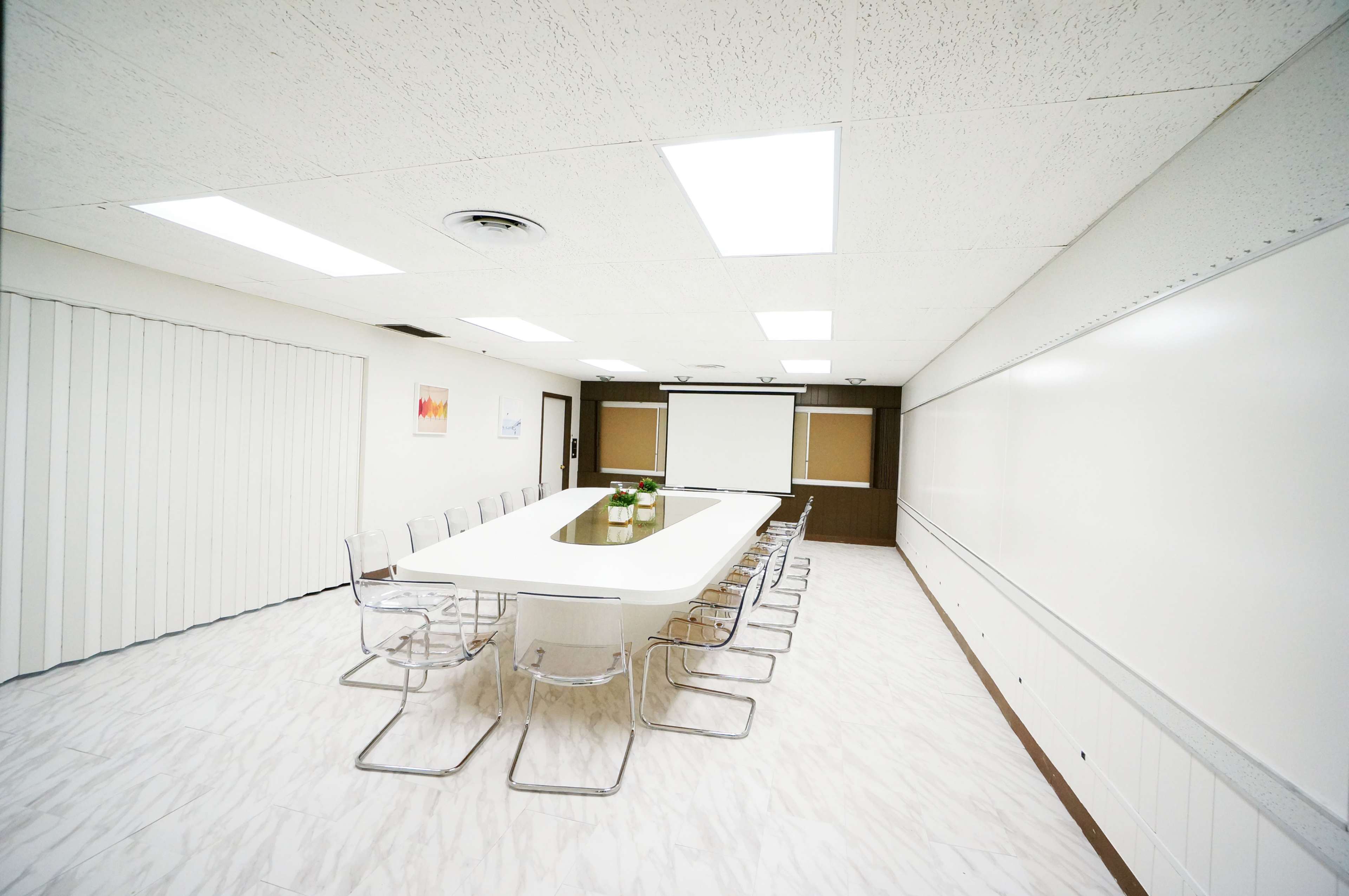 A large conference room features a long white table surrounded by clear chairs, with a projector screen and whiteboard on one wall.