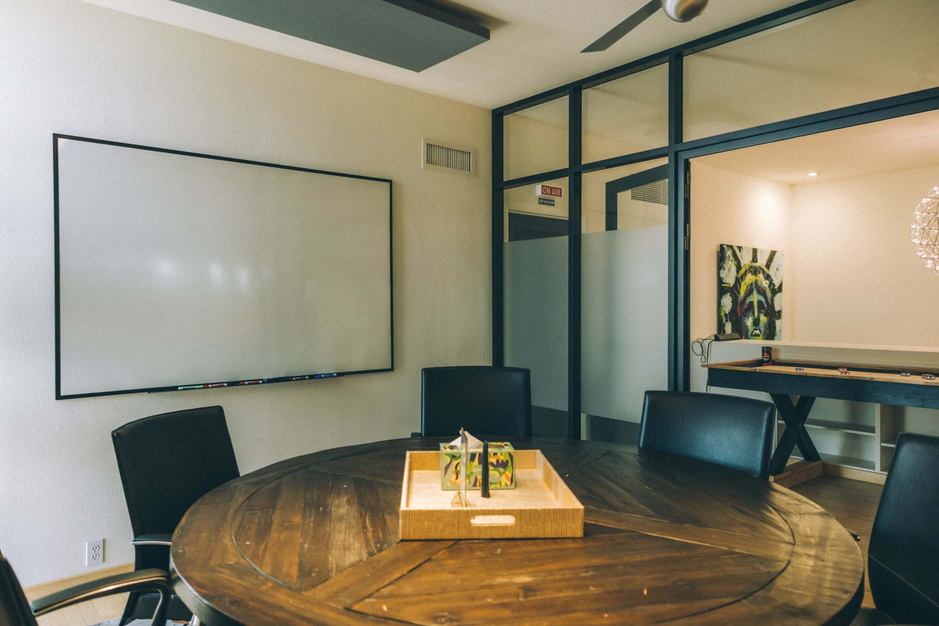 A conference room features a round wooden table surrounded by black chairs, with a large whiteboard on one wall and a glass-walled area in the background.