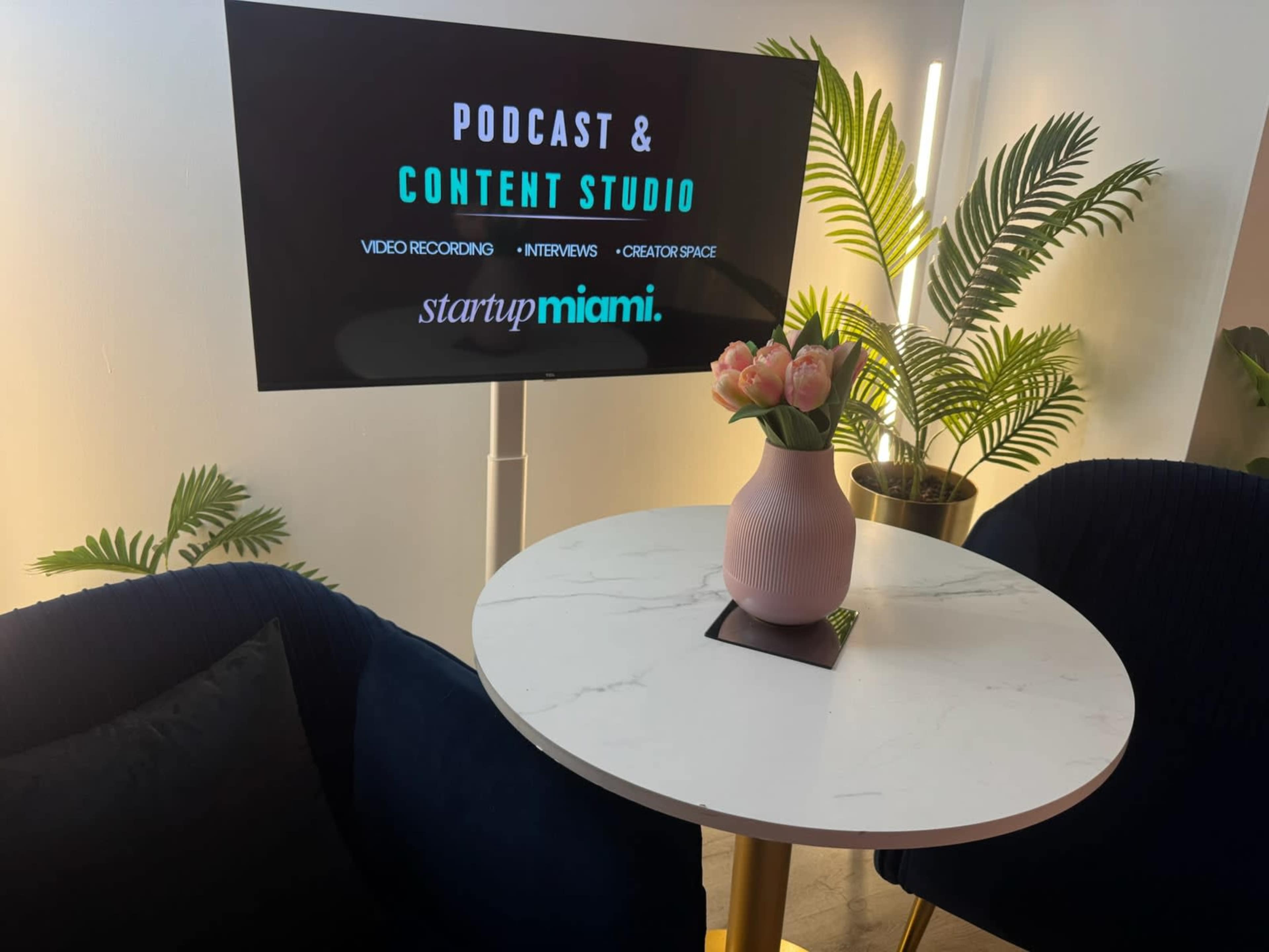 The image shows a modern podcast and content studio featuring a small round table with a vase of tulips, two dark blue chairs, and a television displaying information about the studio.