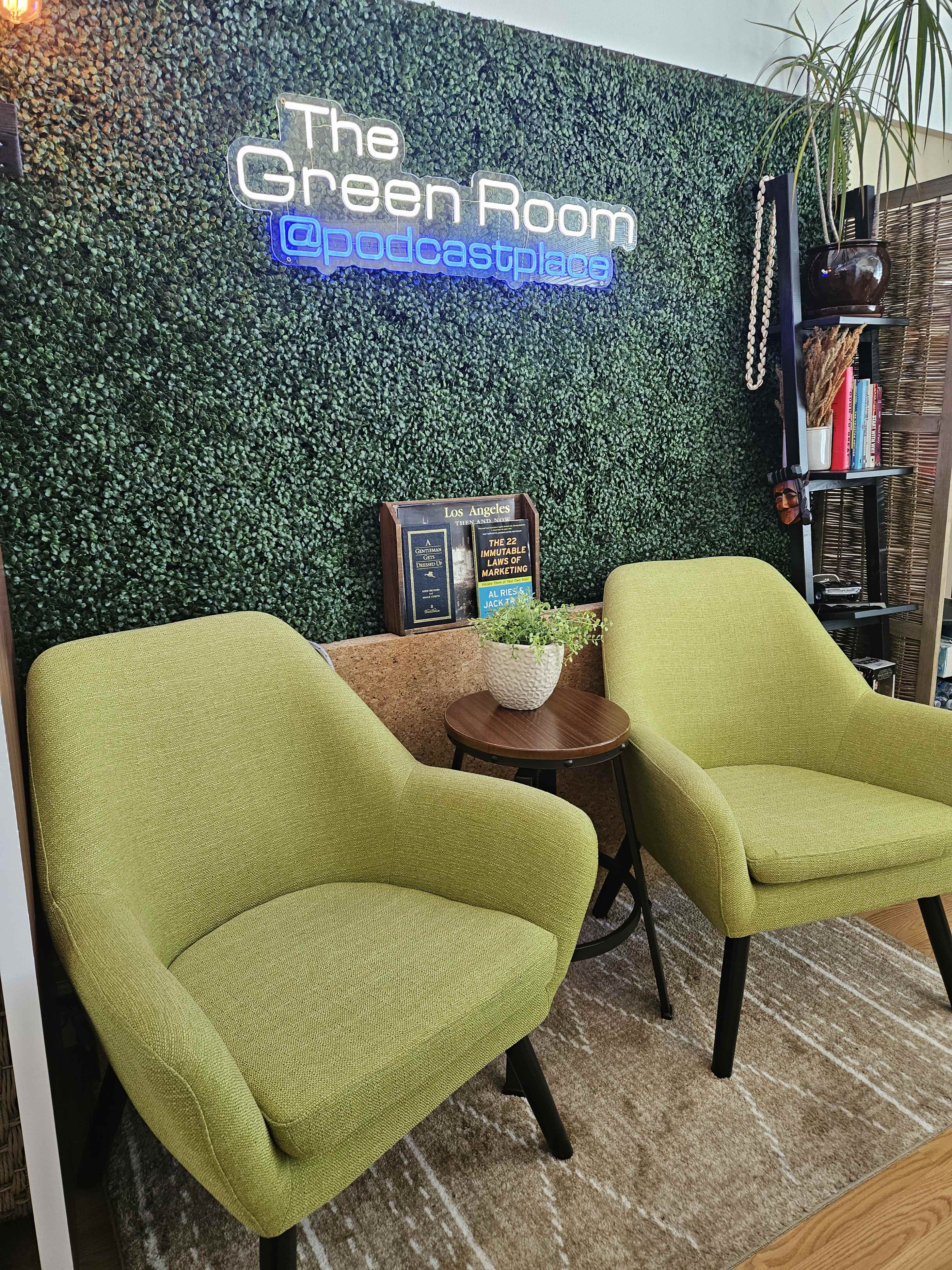 A modern green-themed room with two green chairs, a decorative plant, and a wall featuring the text "The Green Room @podcastplace" in neon lights.