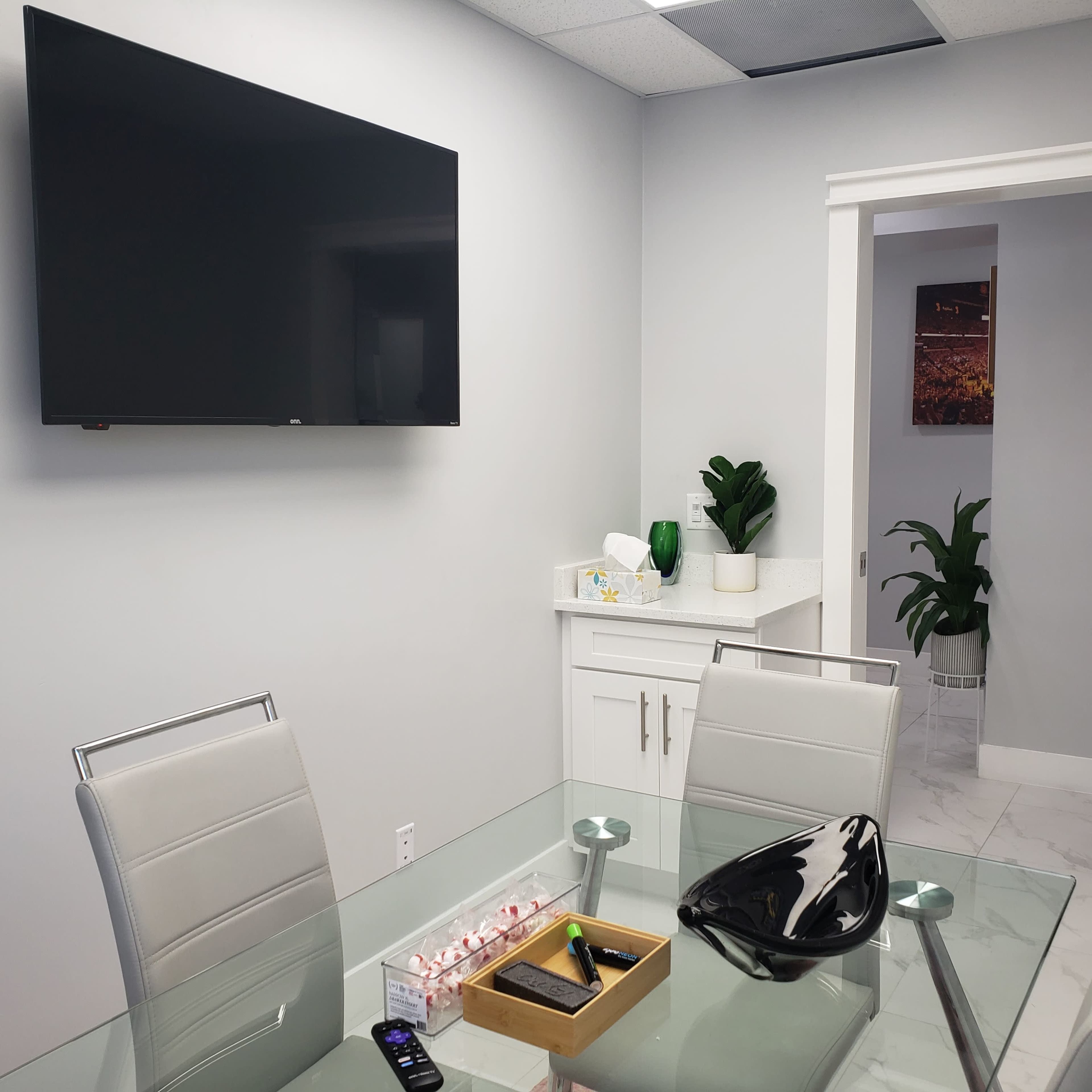 A modern conference room features a glass table, two chairs, a wall-mounted television, and a countertop with decorative items and a plant.