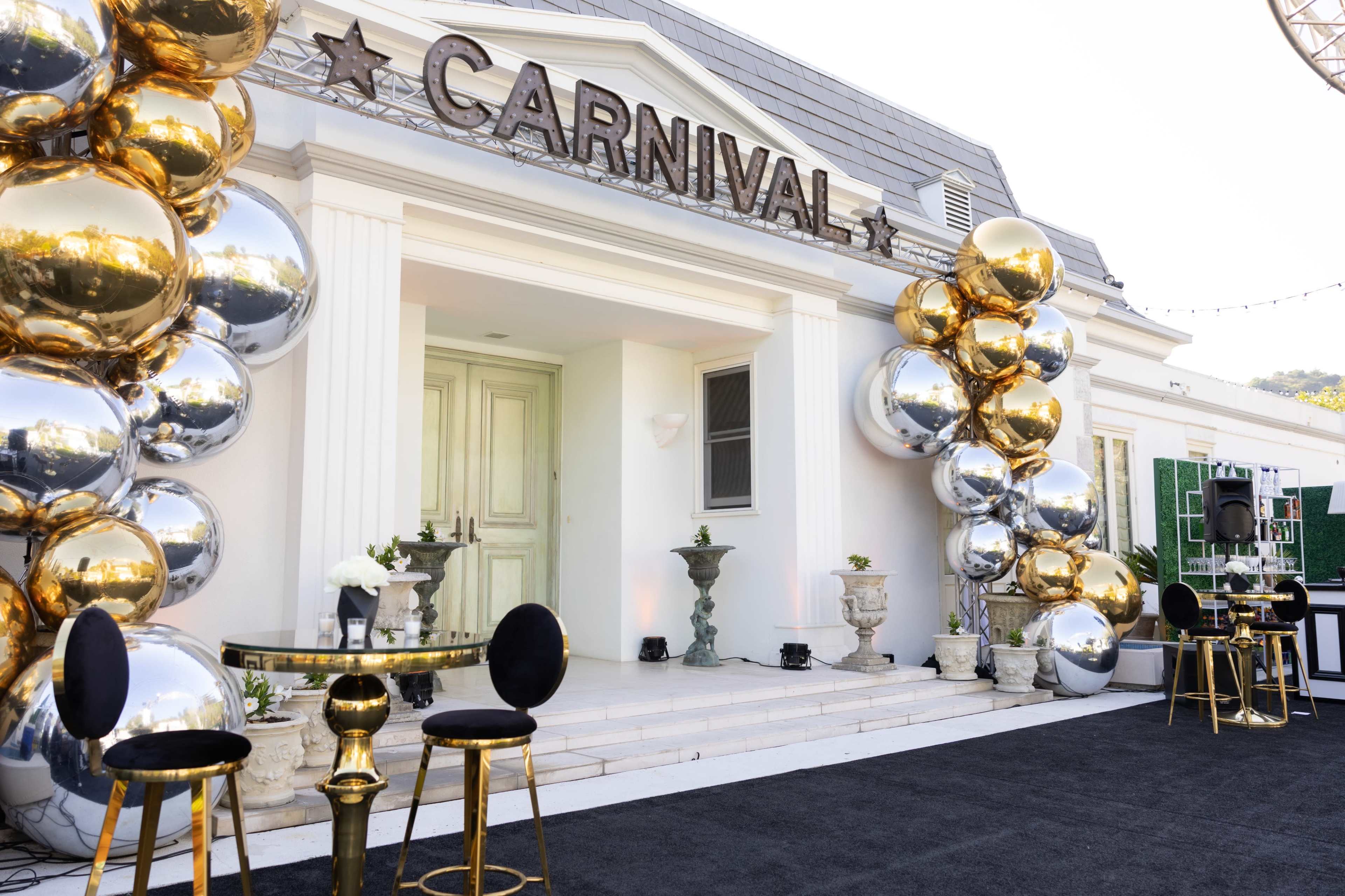 A decorative entrance features a "CARNIVAL" sign, gold and silver balloon arrangements, and elegant black and gold furniture.