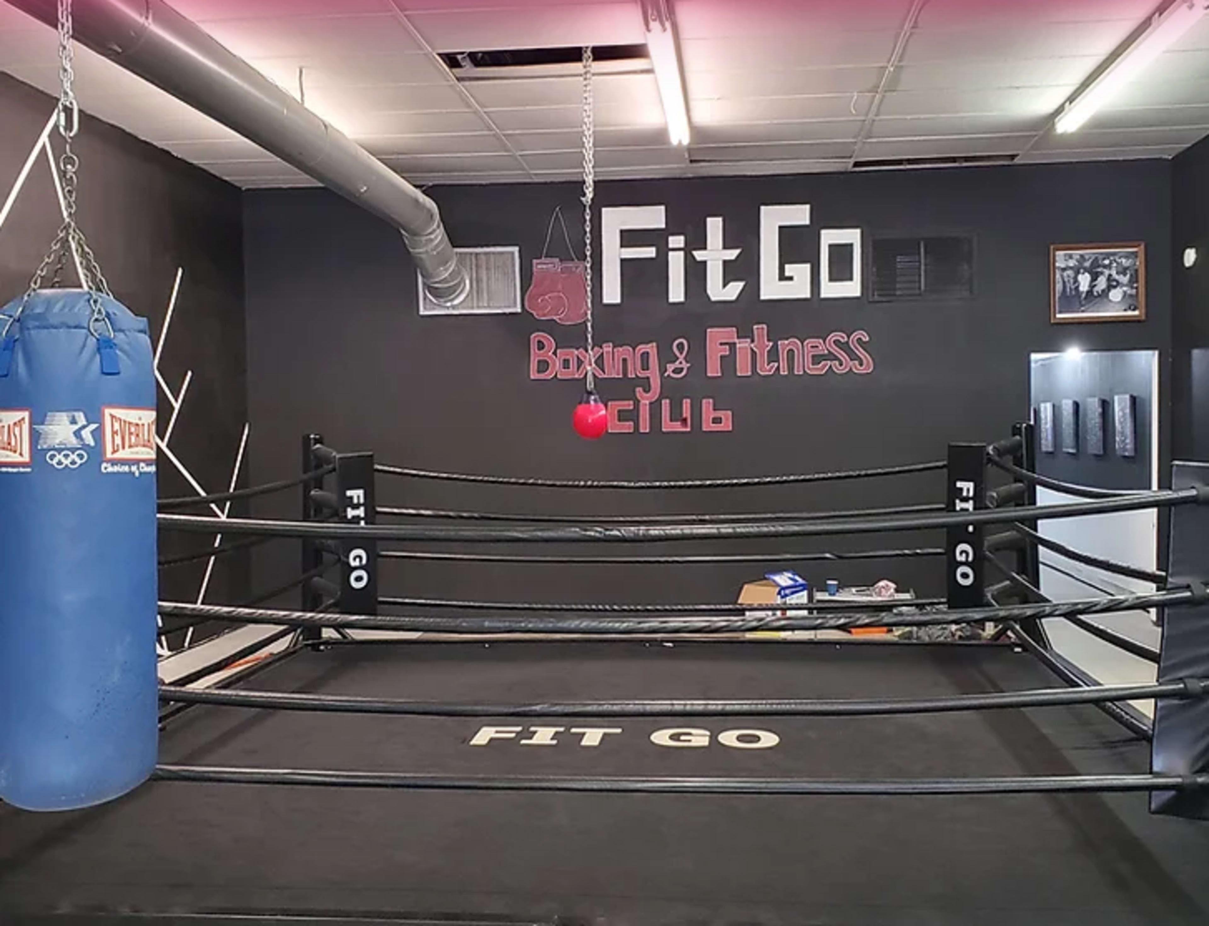 A boxing gym featuring a blue heavy bag and a boxing ring with a black floor, labeled "FIT GO" and "Boxing & Fitness Club" on the walls.