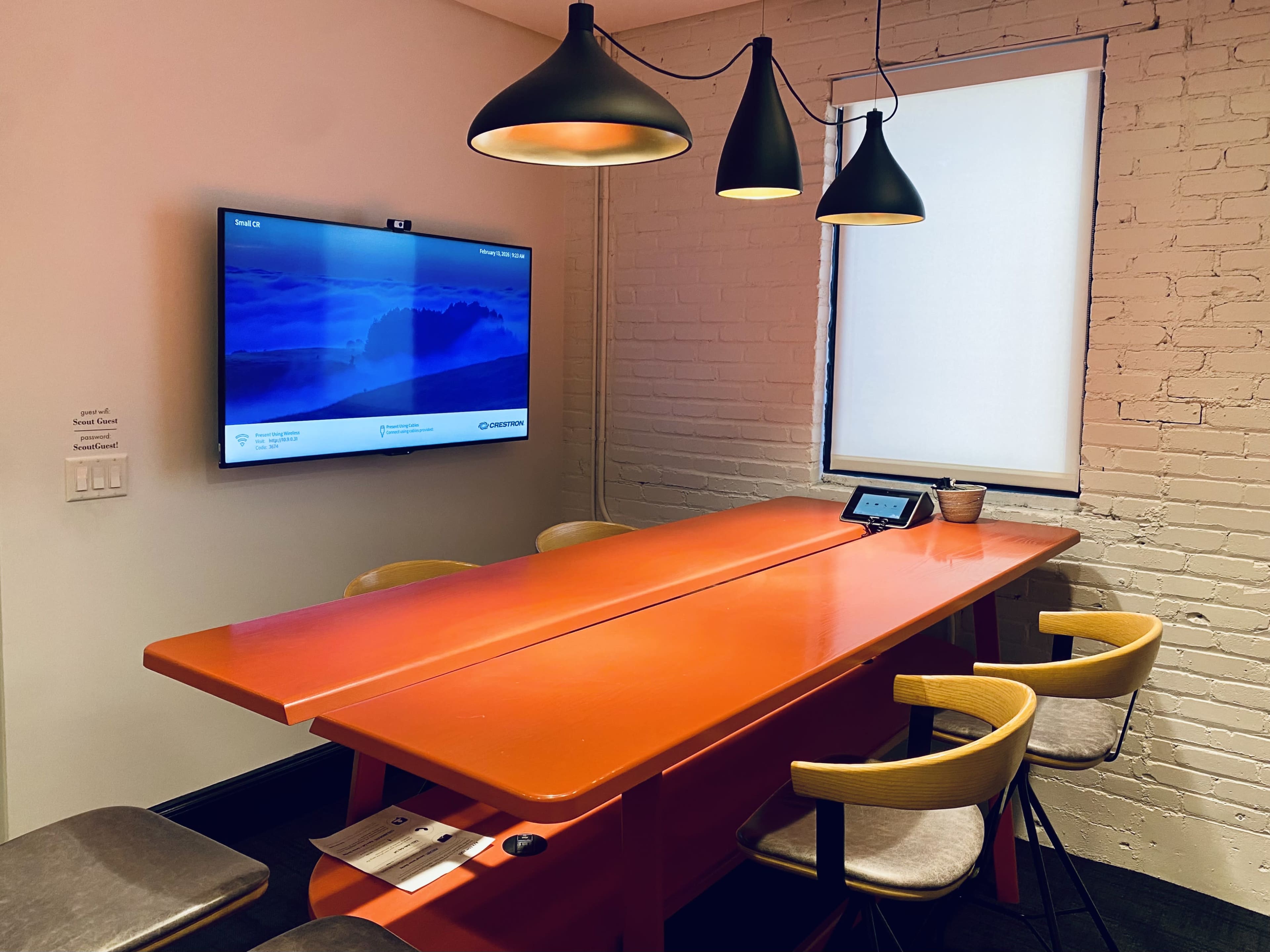 A brightly colored orange table with several chairs is set in a meeting room, featuring a large wall-mounted screen and pendant lighting.