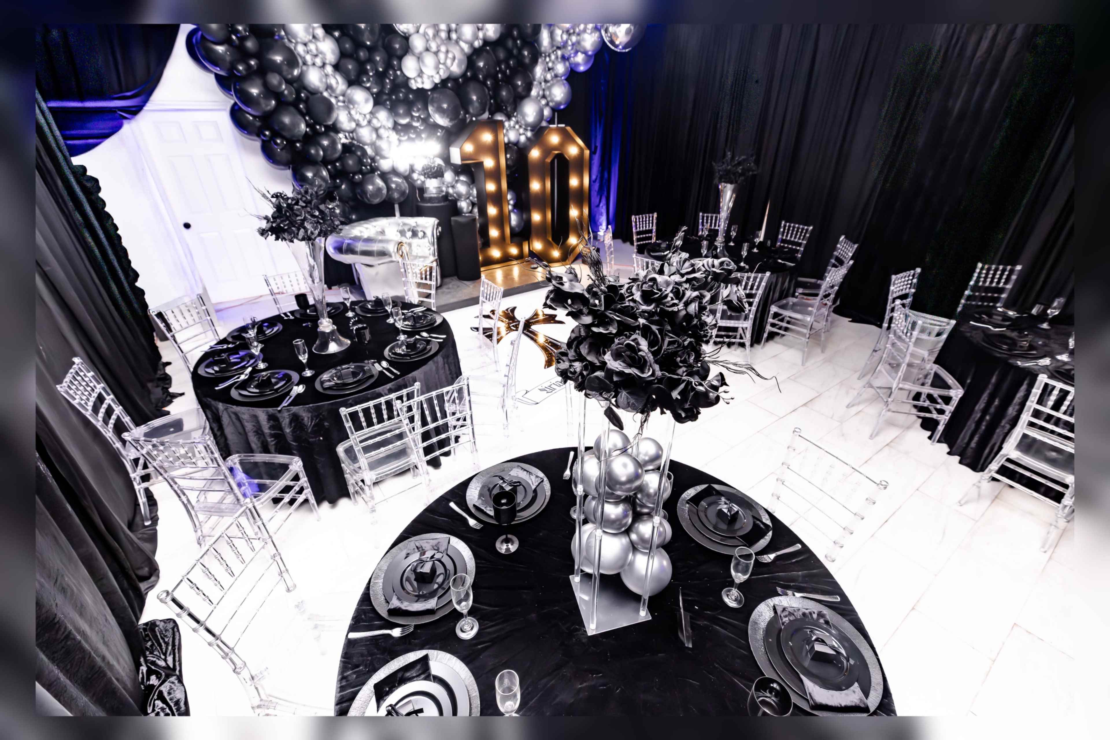The image shows a decorated event space featuring black tables set with elegant dishware, a large "10" made of lighted numbers, and a backdrop of balloons and black drapes.