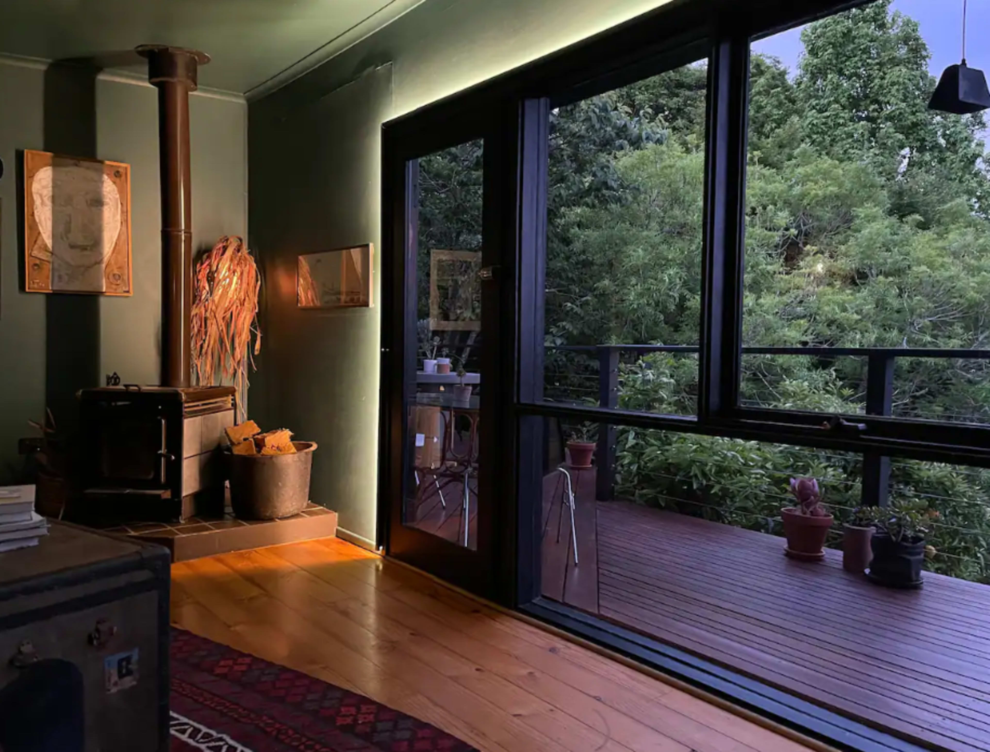 A cozy interior space features a wood stove and large glass doors opening to a deck with lush foliage outside.
