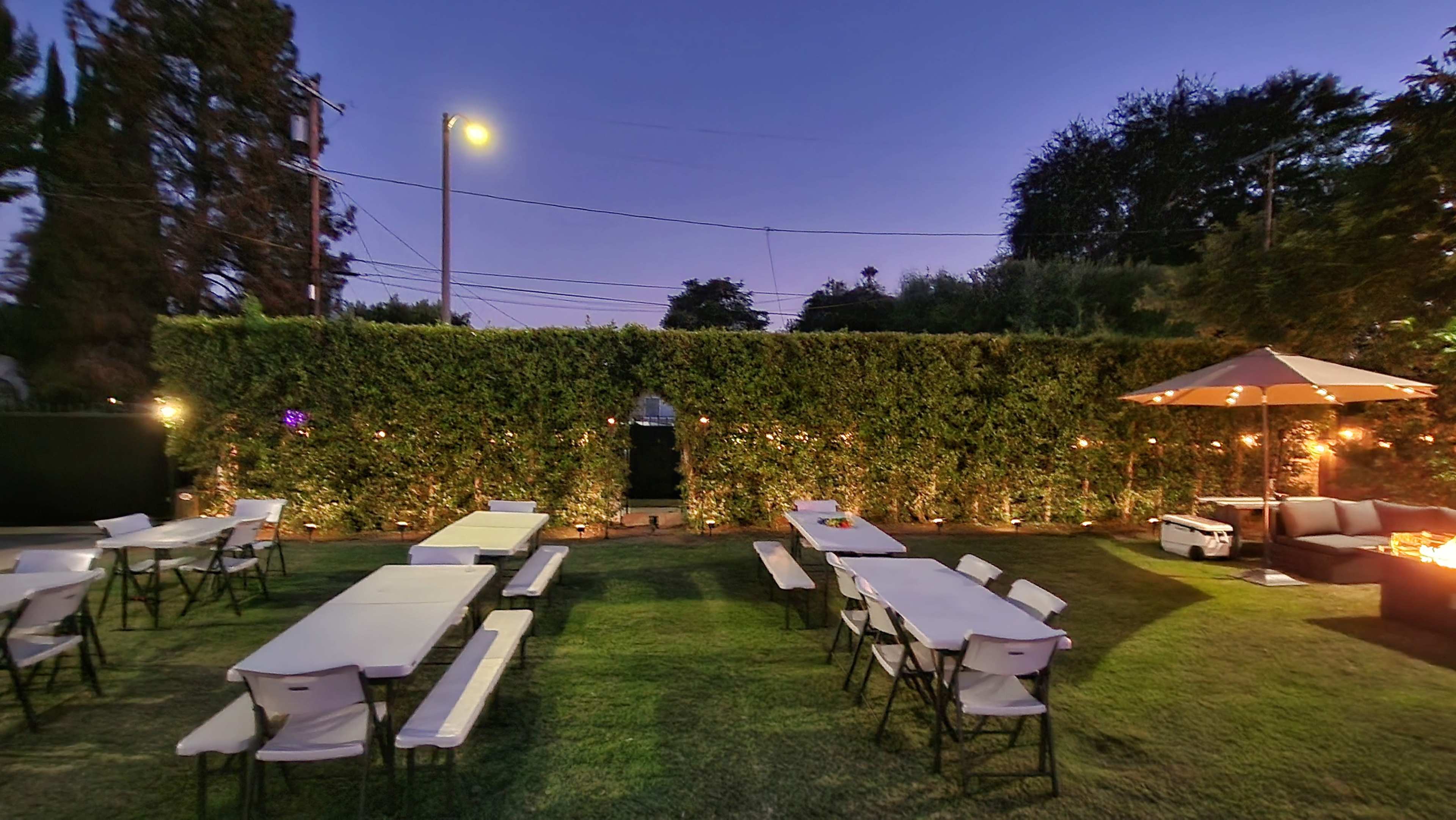The image shows a backyard setting with several white tables and benches arranged on a green lawn, surrounded by a leafy hedge and illuminated by evening lights.