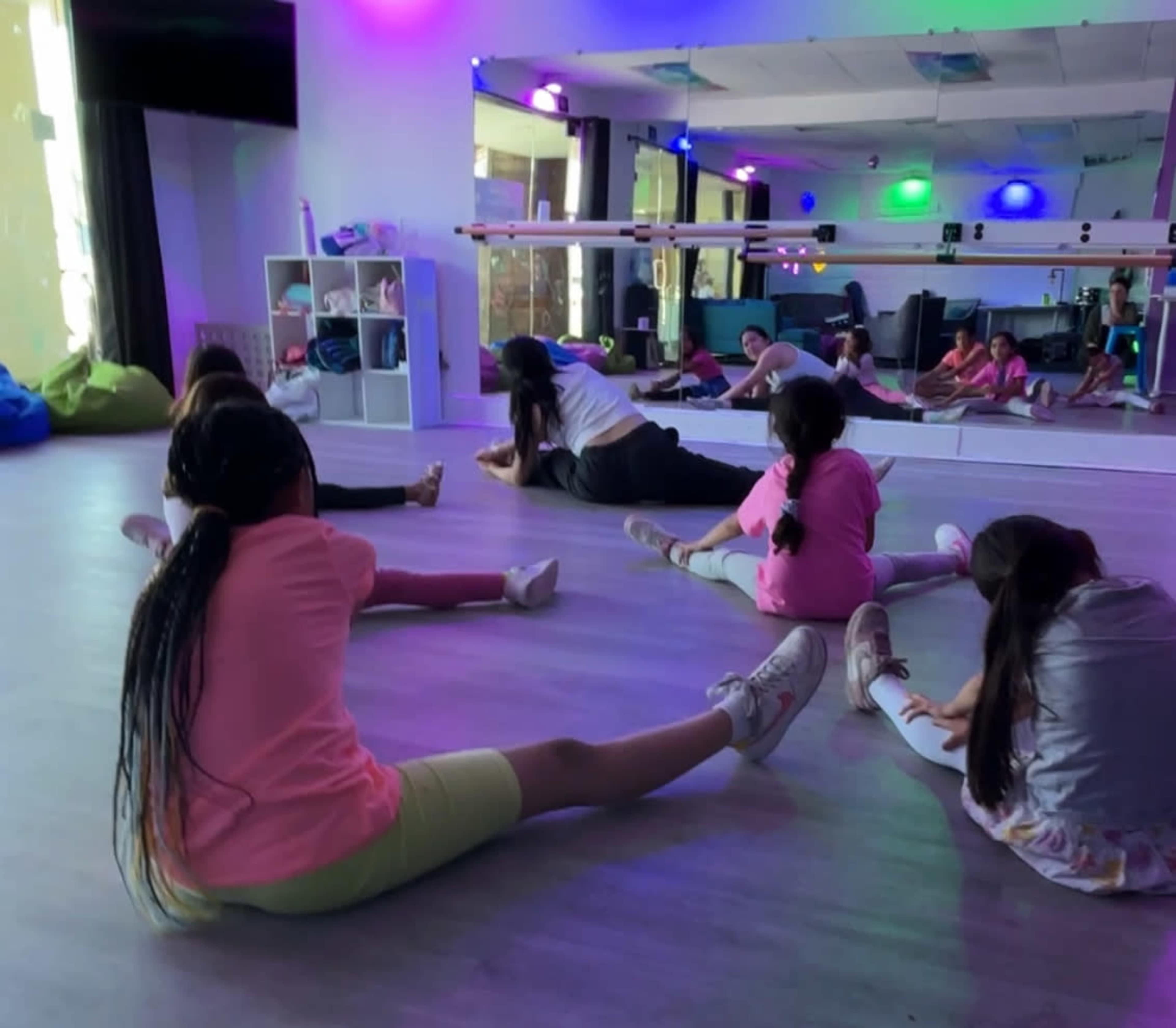 A group of children is stretching on a hardwood floor in a brightly lit dance studio with mirrors and colorful lights.