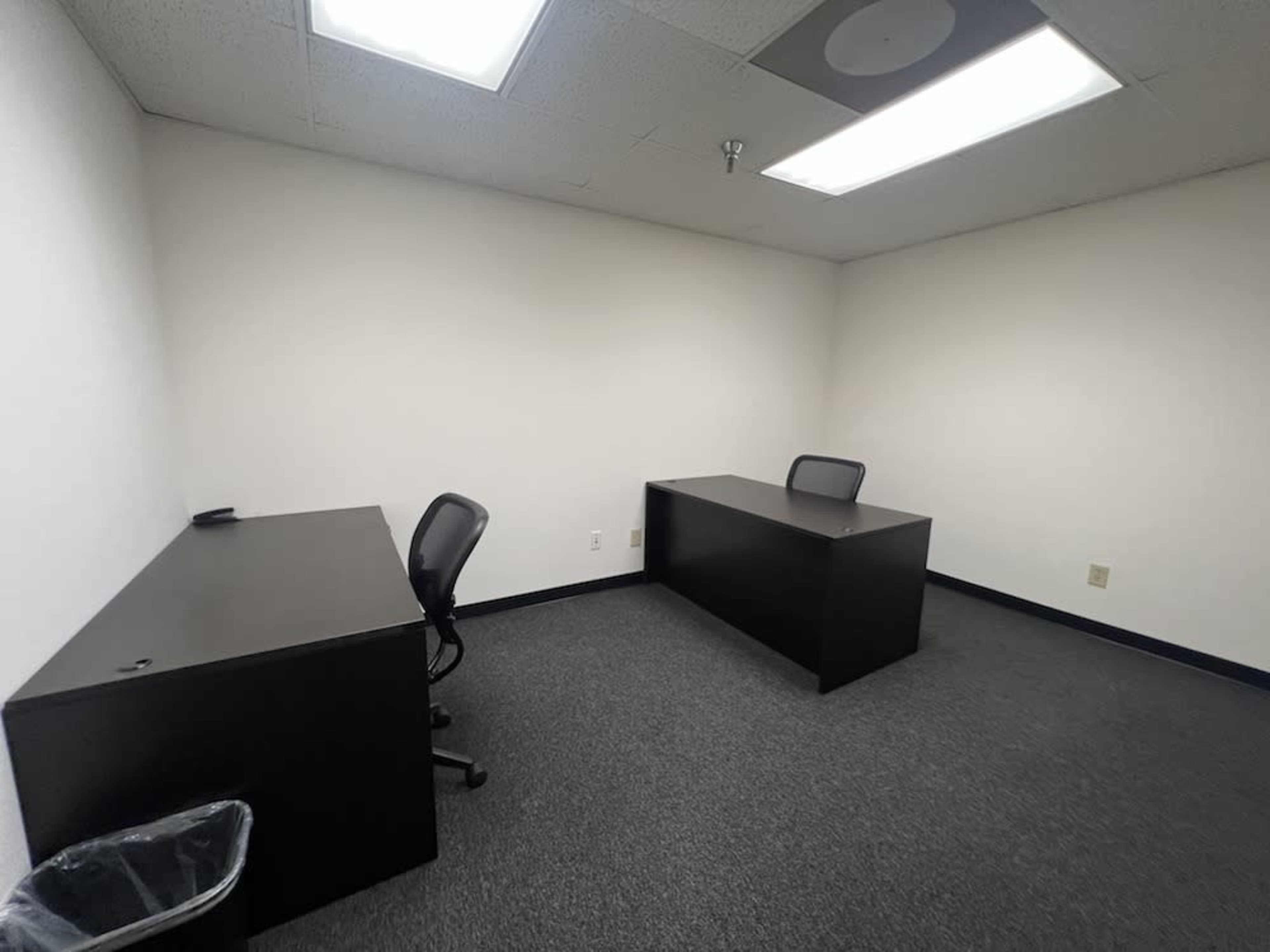 The image shows a bare office space featuring two dark-colored desks with chairs and a trash can in the corner.