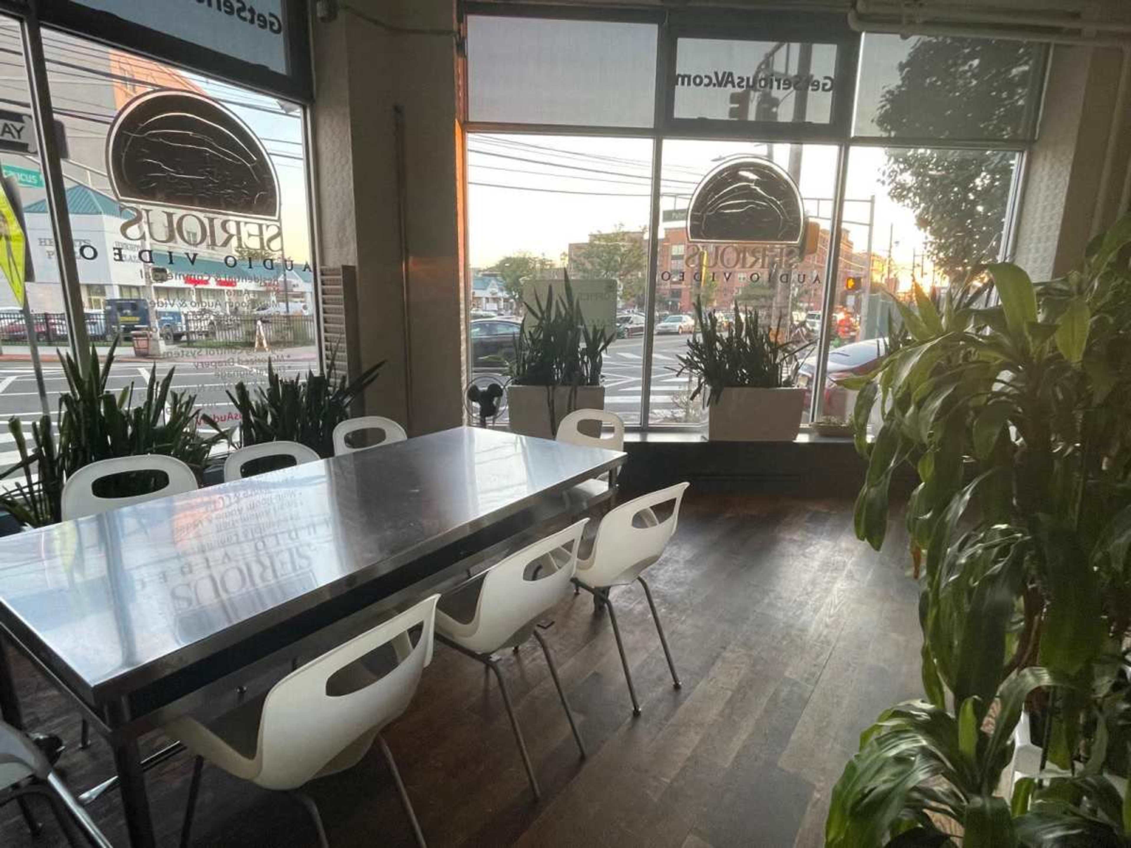 A modern dining area features a large table surrounded by white chairs, with floor-to-ceiling windows showcasing an outdoor view at sunset.