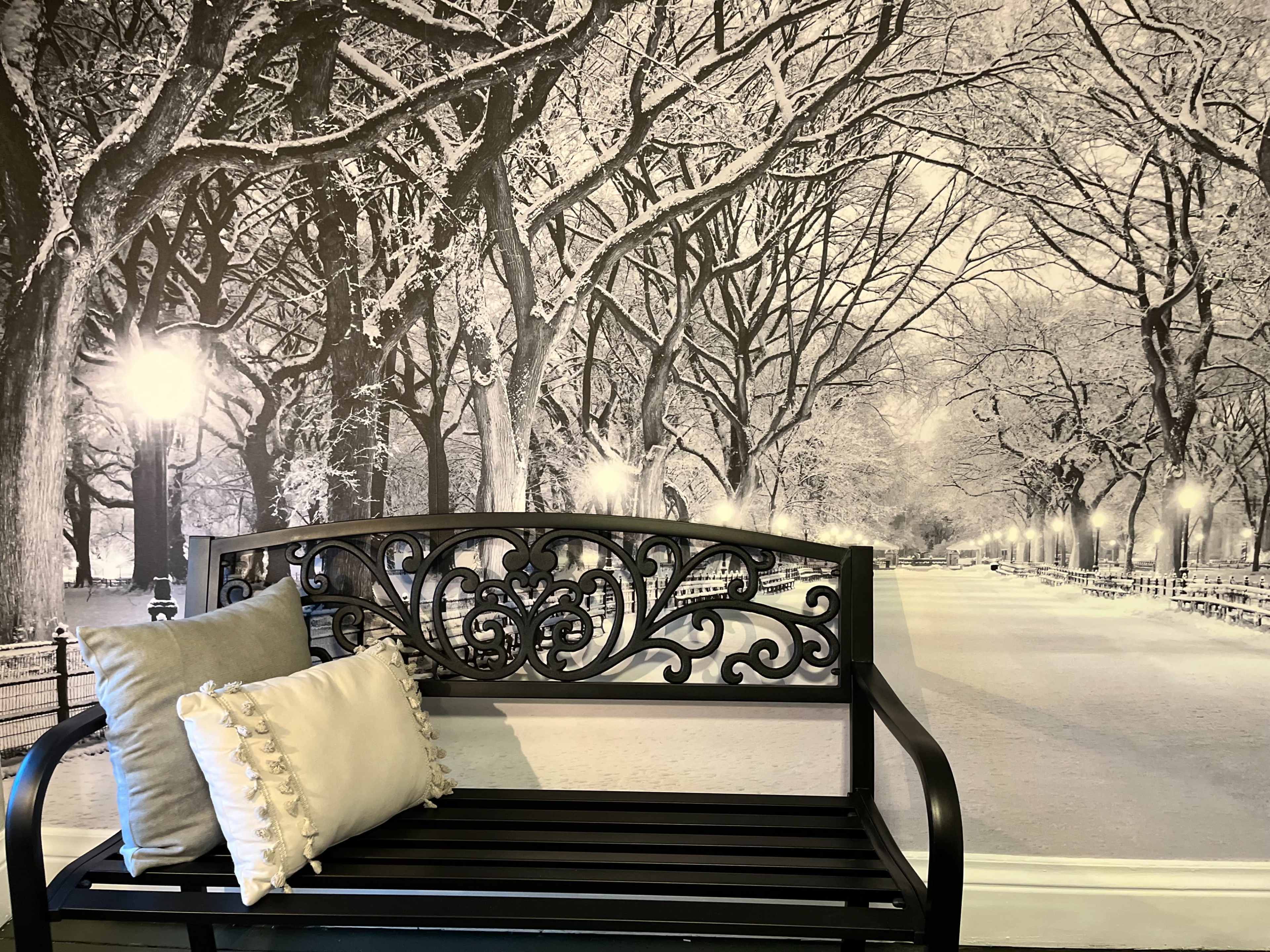 A black metal bench with decorative details is positioned in front of a black-and-white mural depicting a snow-covered park scene with trees and lights.