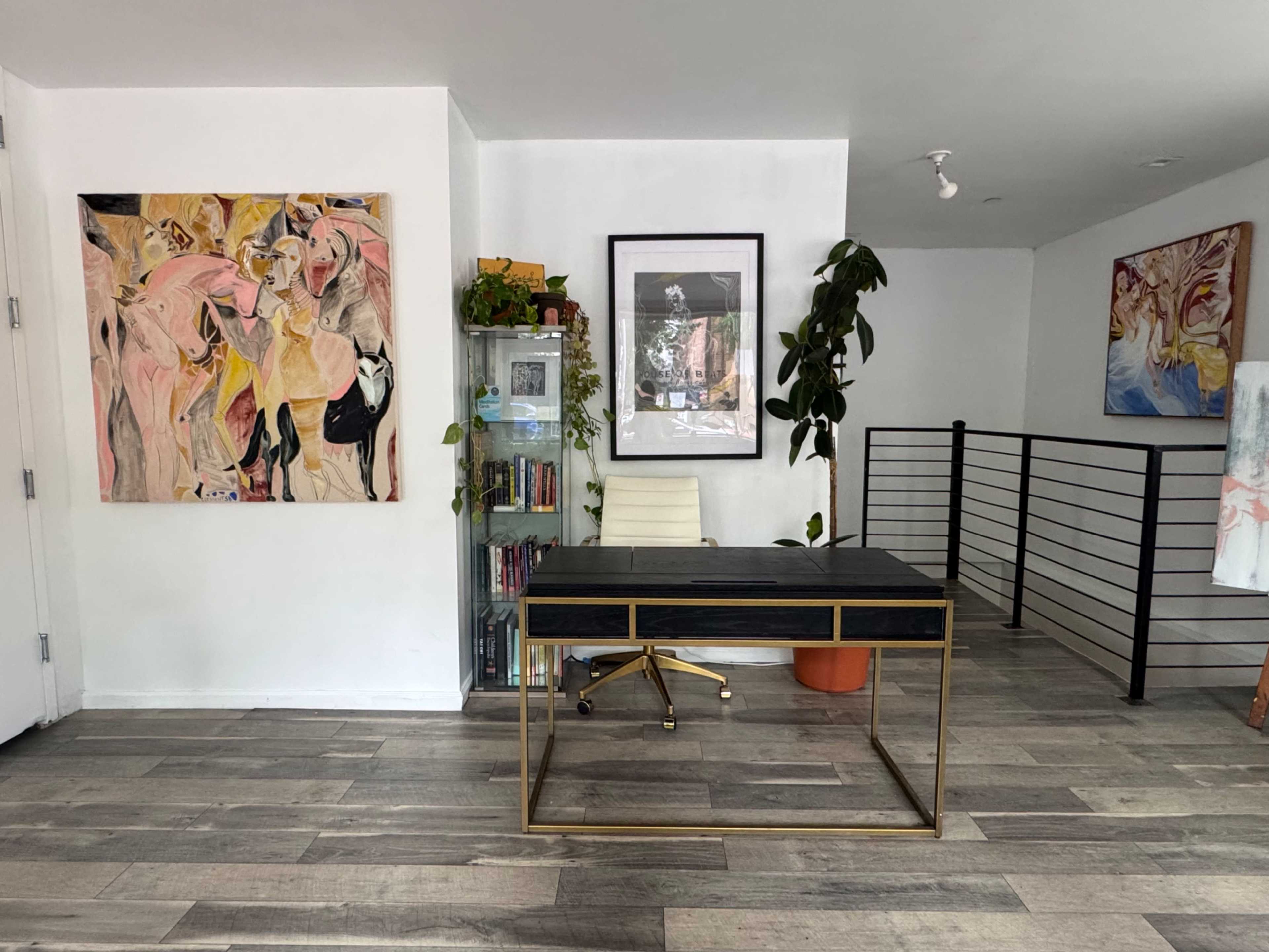 The image shows a modern workspace featuring a desk with a black top and gold frame, surrounded by art on the walls and various plants placed in the room.