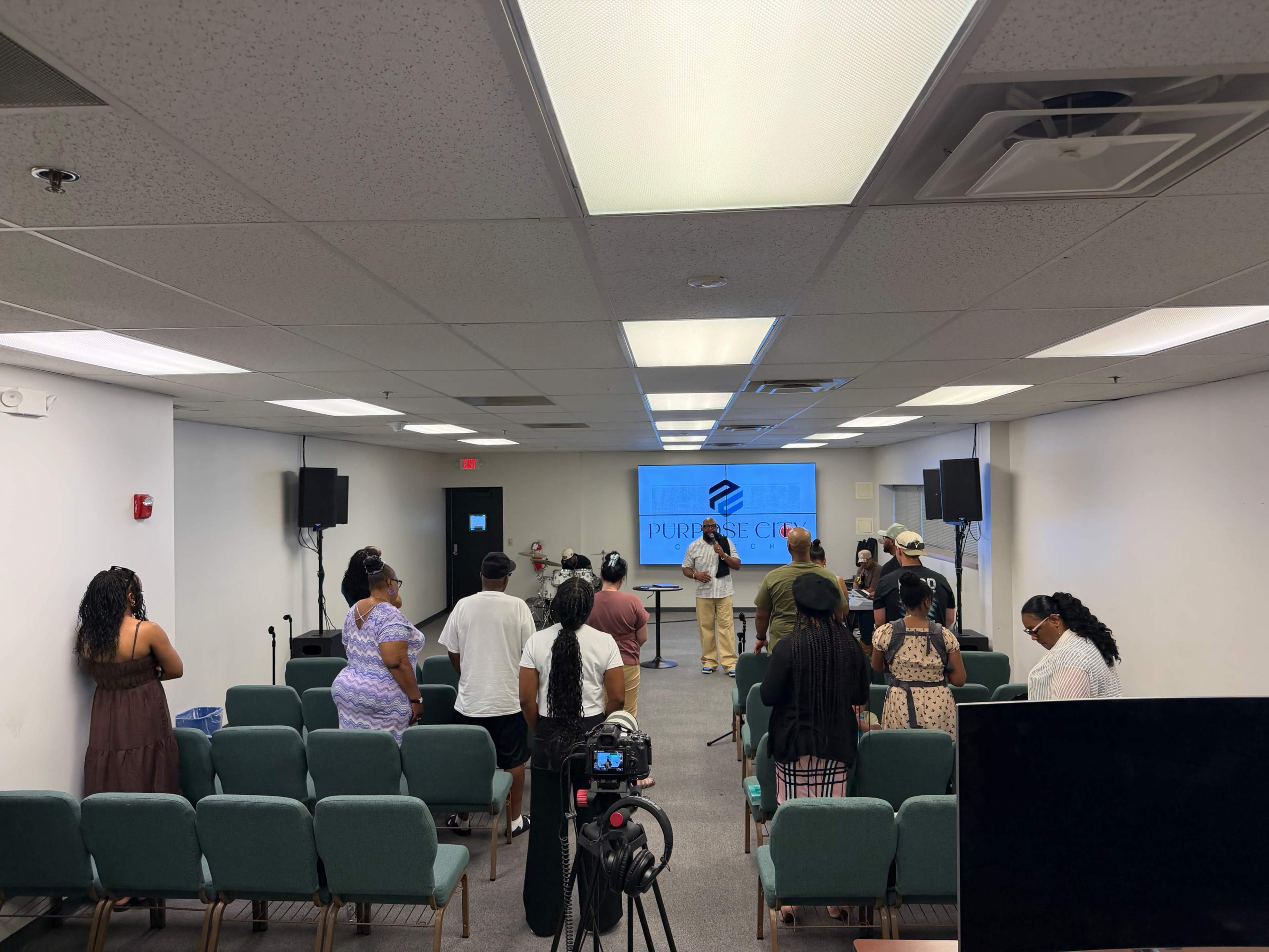 A group of people is gathered in a conference room with seating facing a presentation screen displaying the word "PURPOSE."