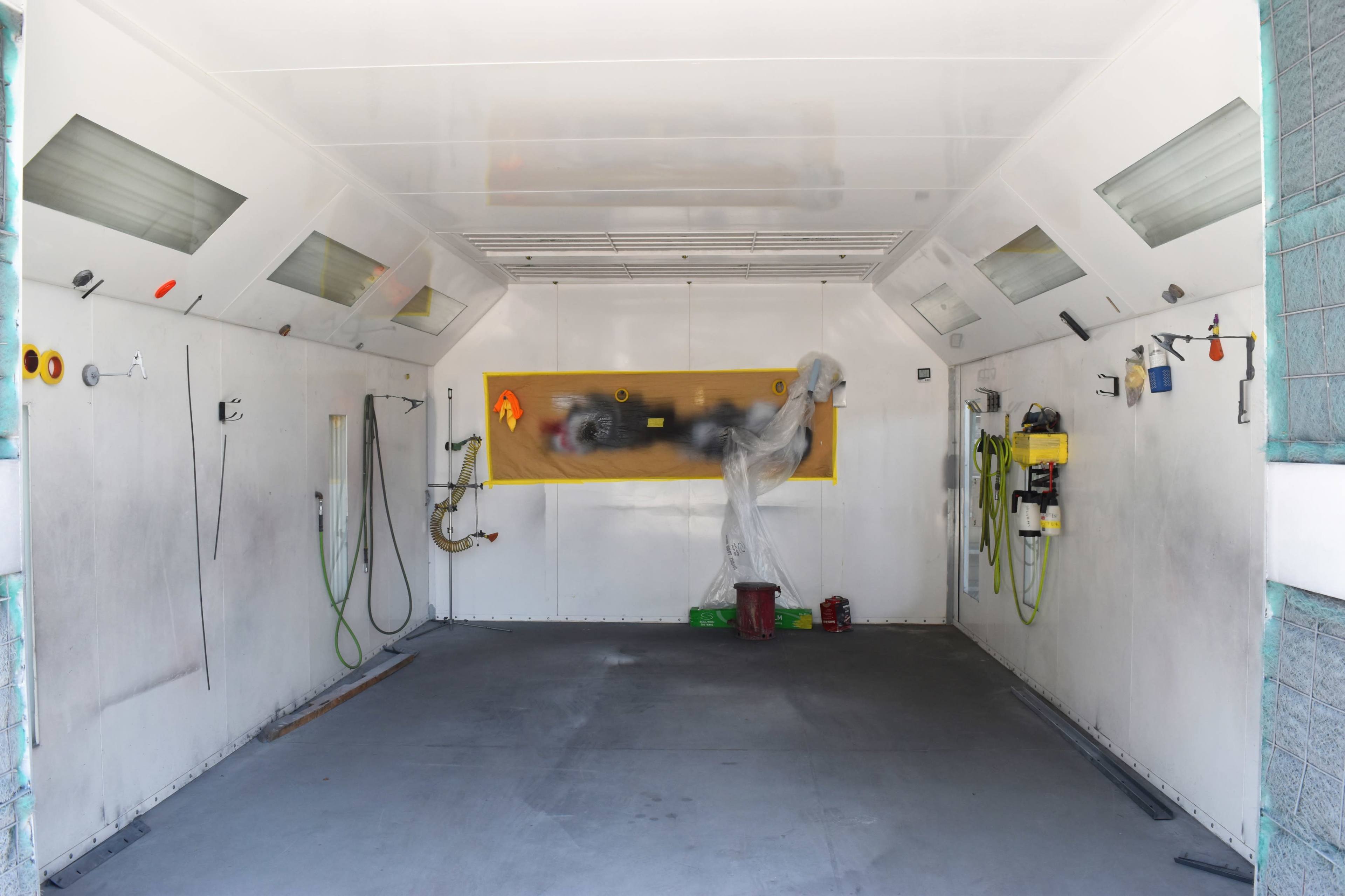 The image shows an empty paint booth with white walls, a smooth floor, and various tools and hoses mounted on the walls.