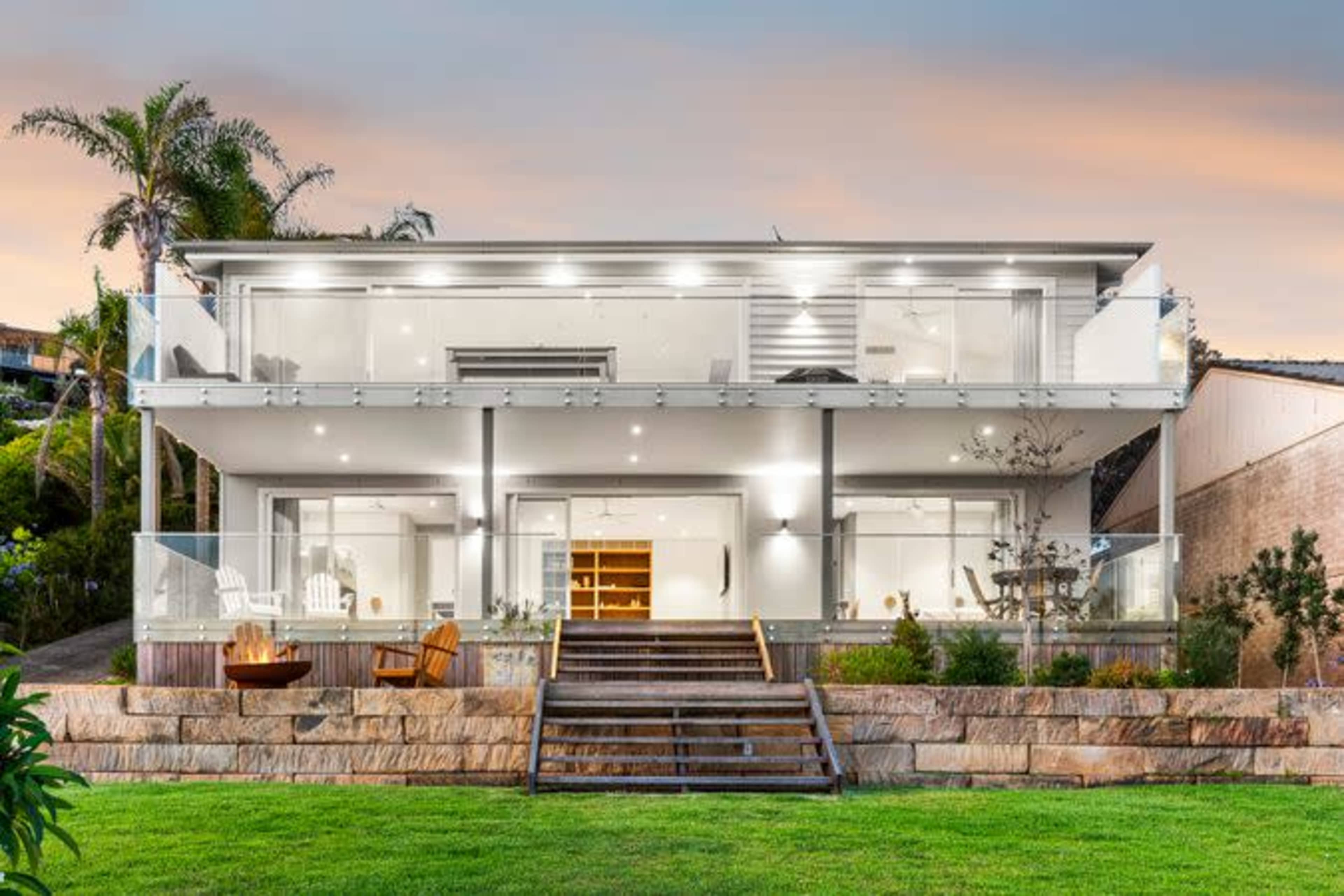 A modern two-story house with large glass balconies, wooden steps leading to a grassy yard, and palm trees in the background.
