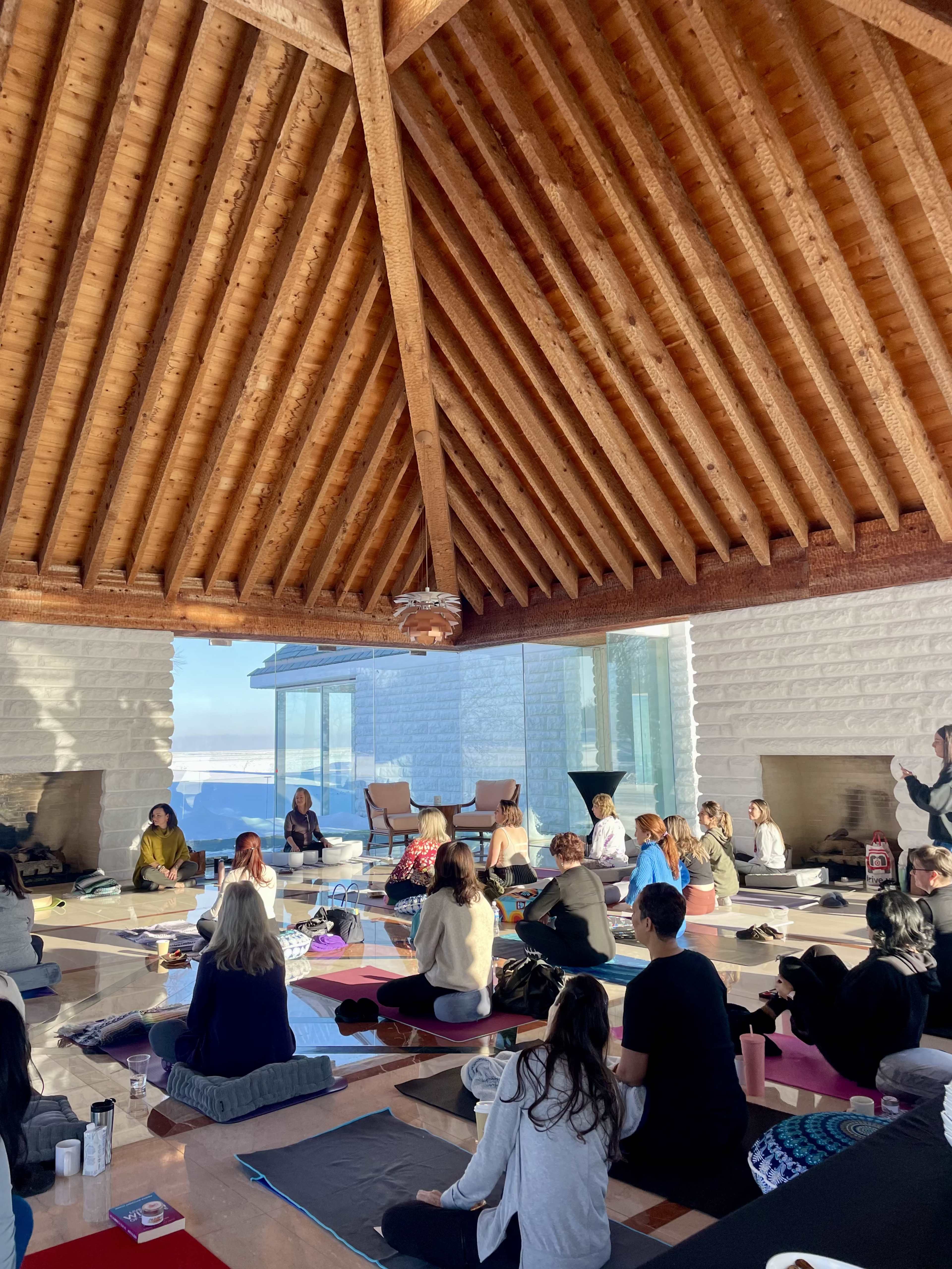 A group of people sit on yoga mats in a spacious, wooden-roofed room with large windows overlooking a scenic view.