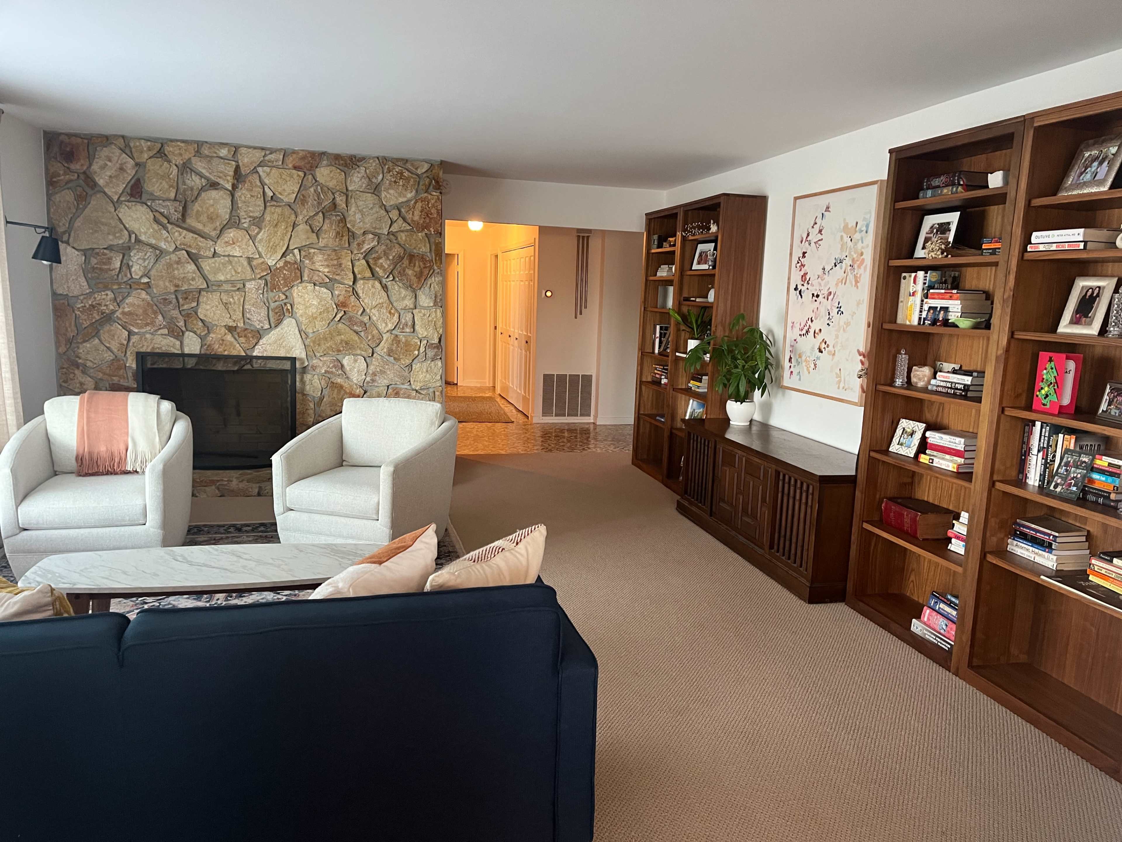 A living room with a stone fireplace, two white armchairs, a dark blue couch, and wooden bookshelves filled with books.