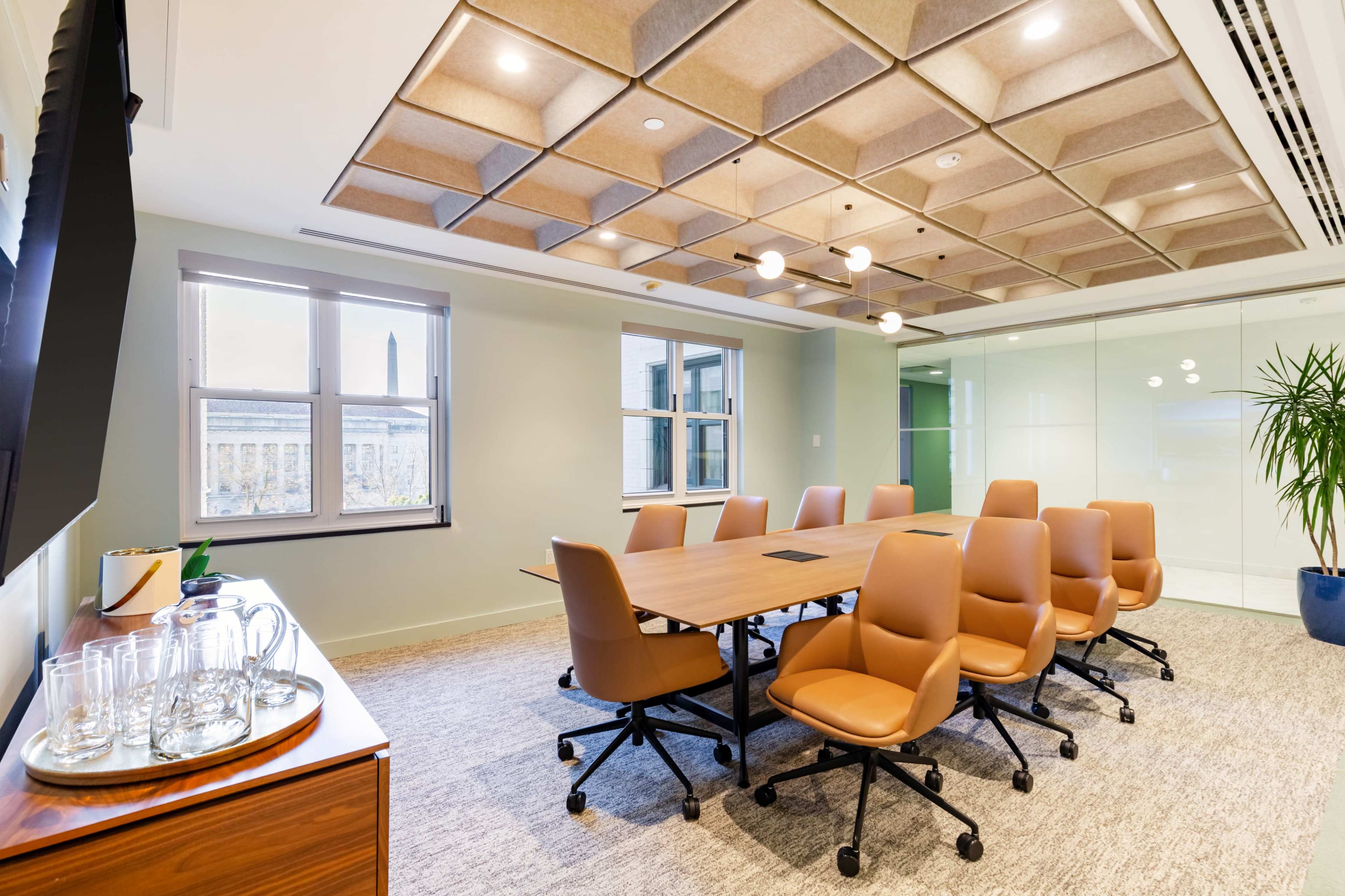 The image shows a modern conference room featuring a large wooden table surrounded by brown ergonomic chairs, with a glass wall and a plant in the corner.