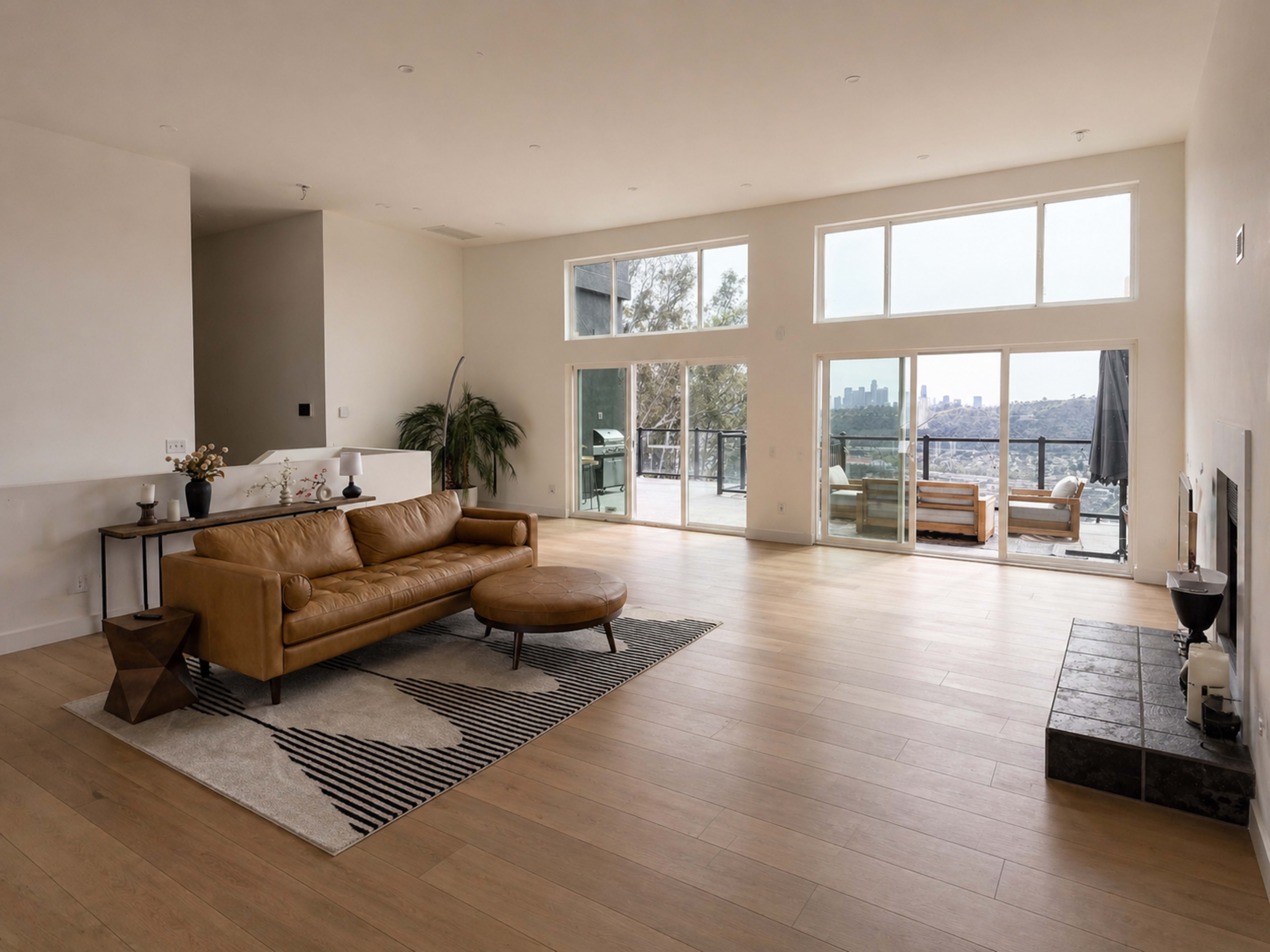 The image shows a spacious living room with a brown leather couch, a round coffee table, and large windows offering a view of the city outside.
