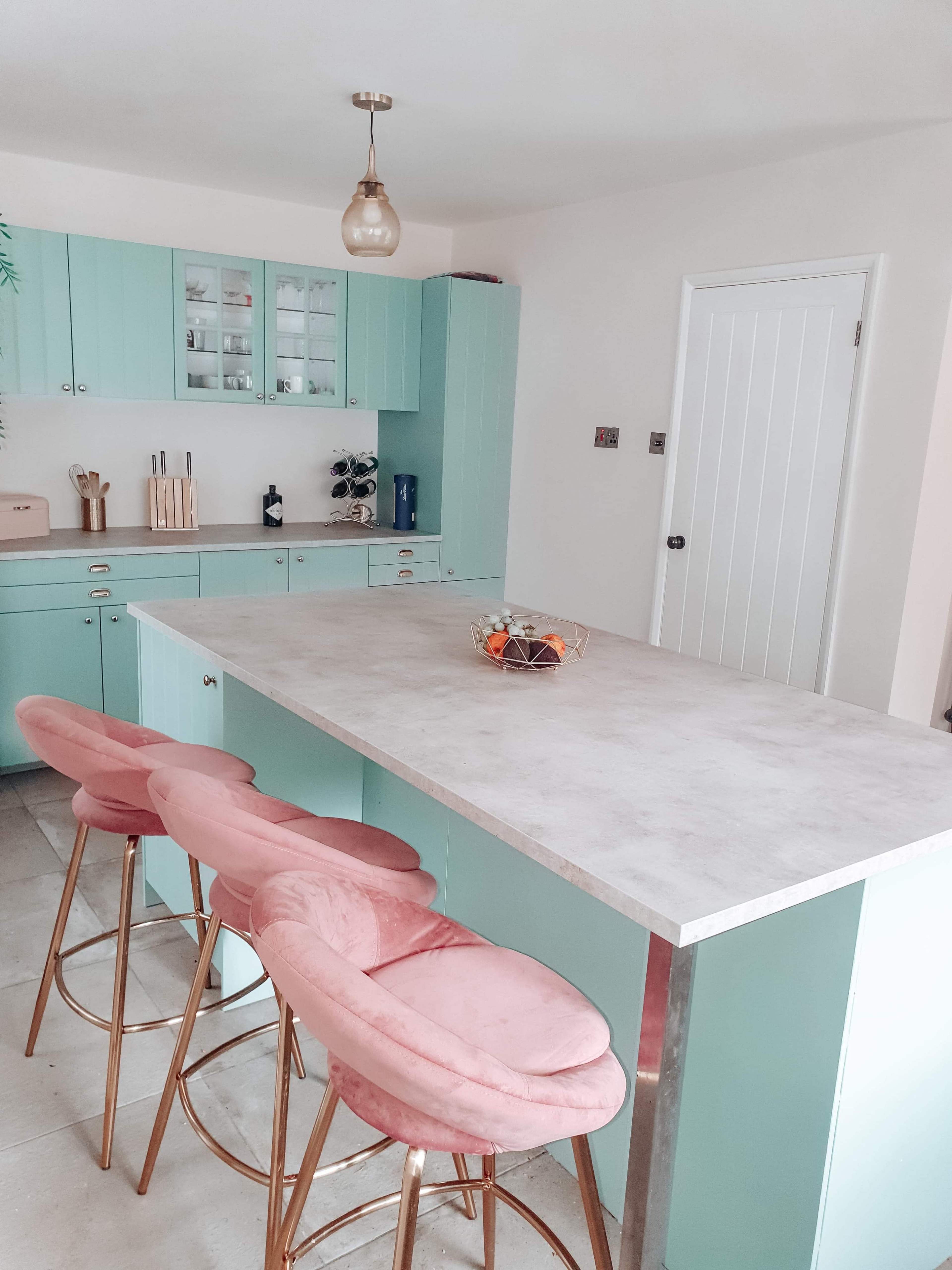 A modern kitchen with mint green cabinetry, a light gray countertop, and pink bar stools arranged around a large island.