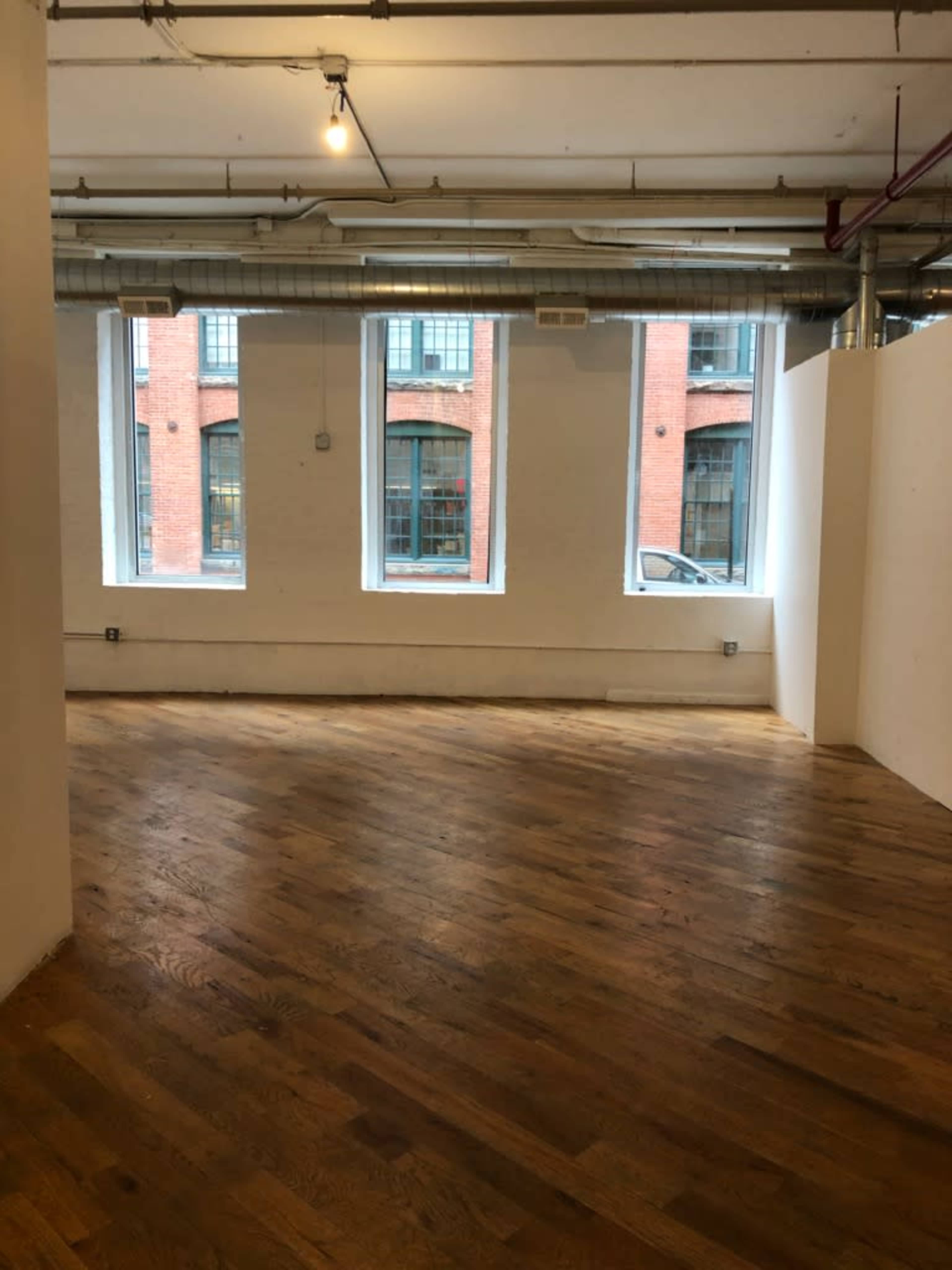 The image shows an empty room with wooden floorboards and large windows that overlook a brick building.