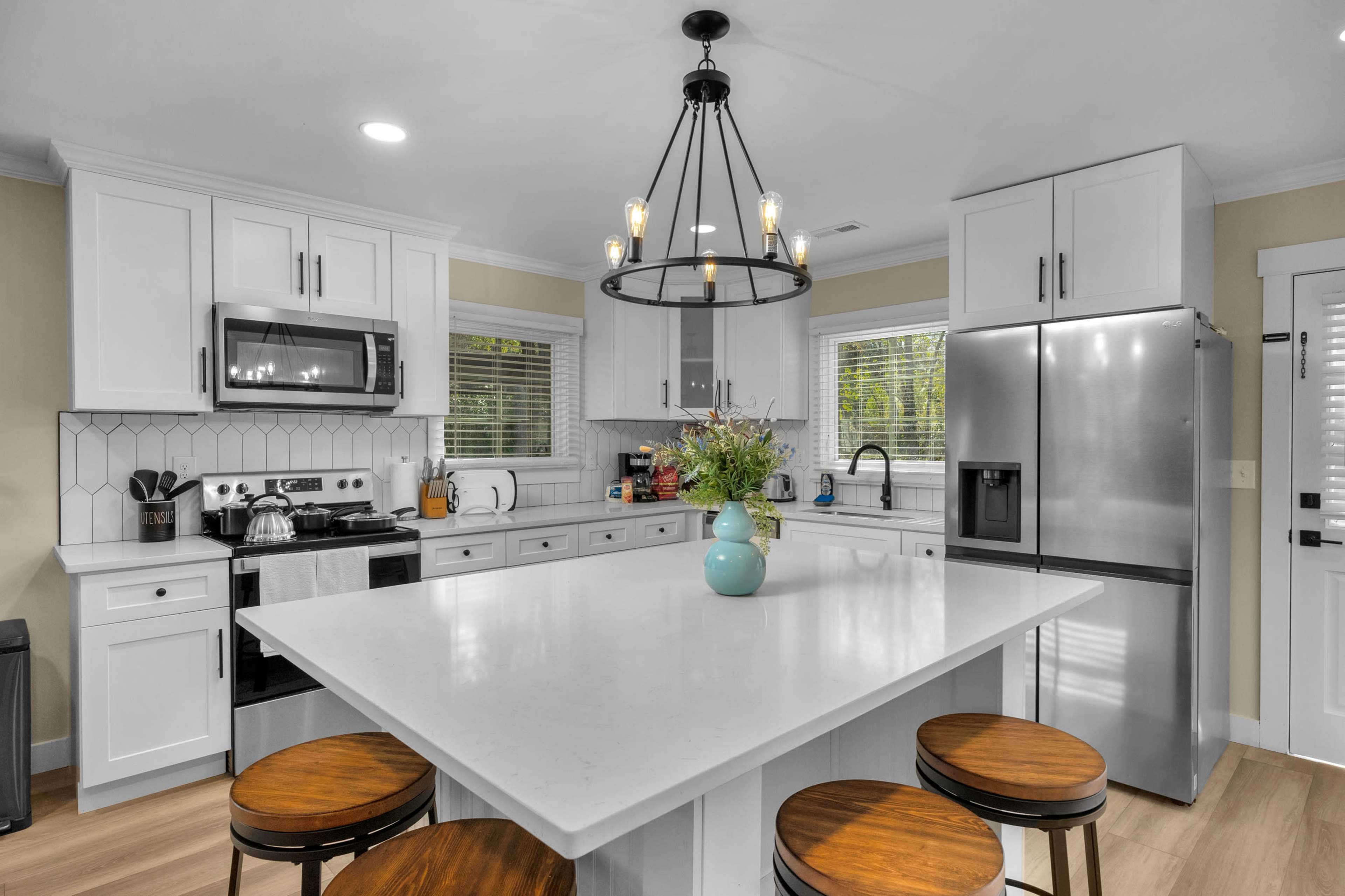 A modern kitchen features a central island with four wooden stools, stainless steel appliances, and ample natural light from two windows.