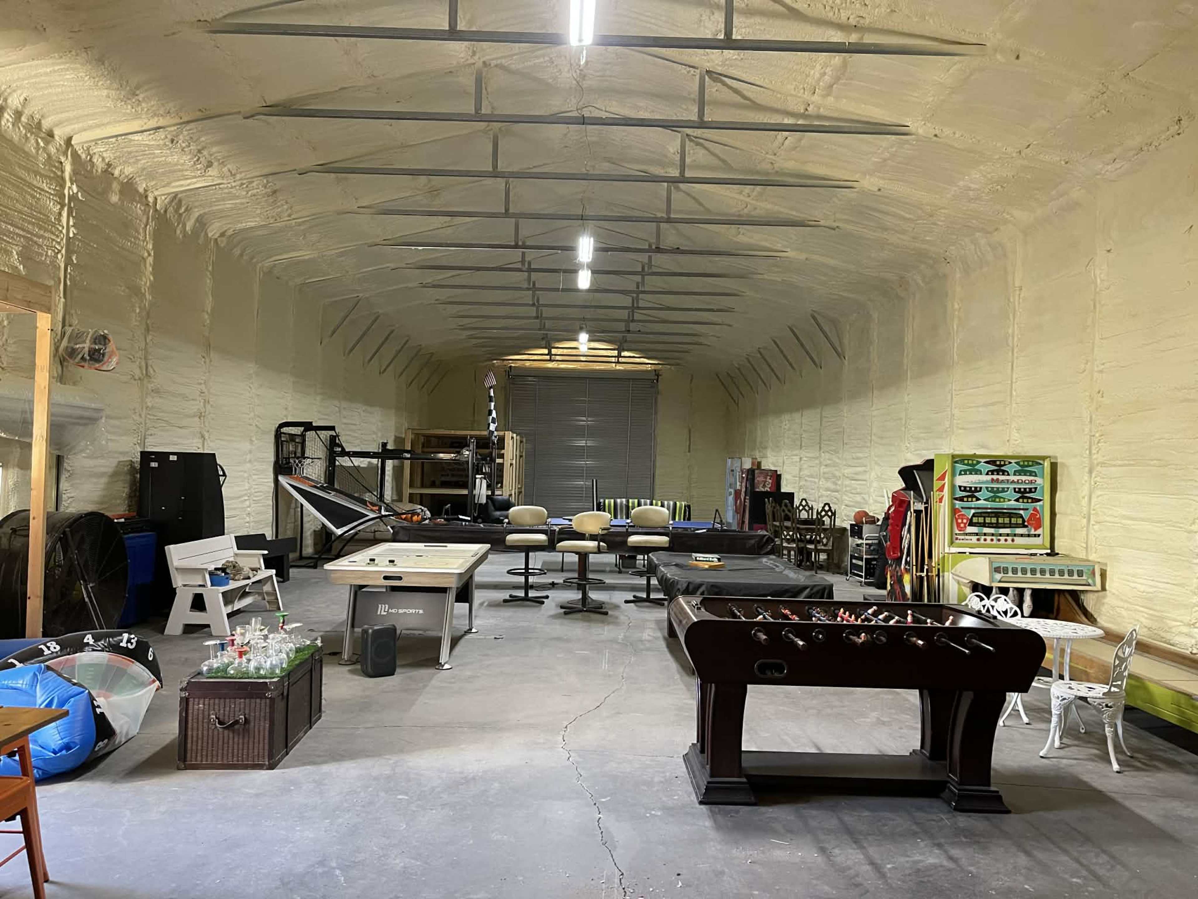The image shows a spacious garage or recreational room featuring various games, including a foosball table, pool table, and arcade game, all surrounded by equipment and furniture against a textured wall.