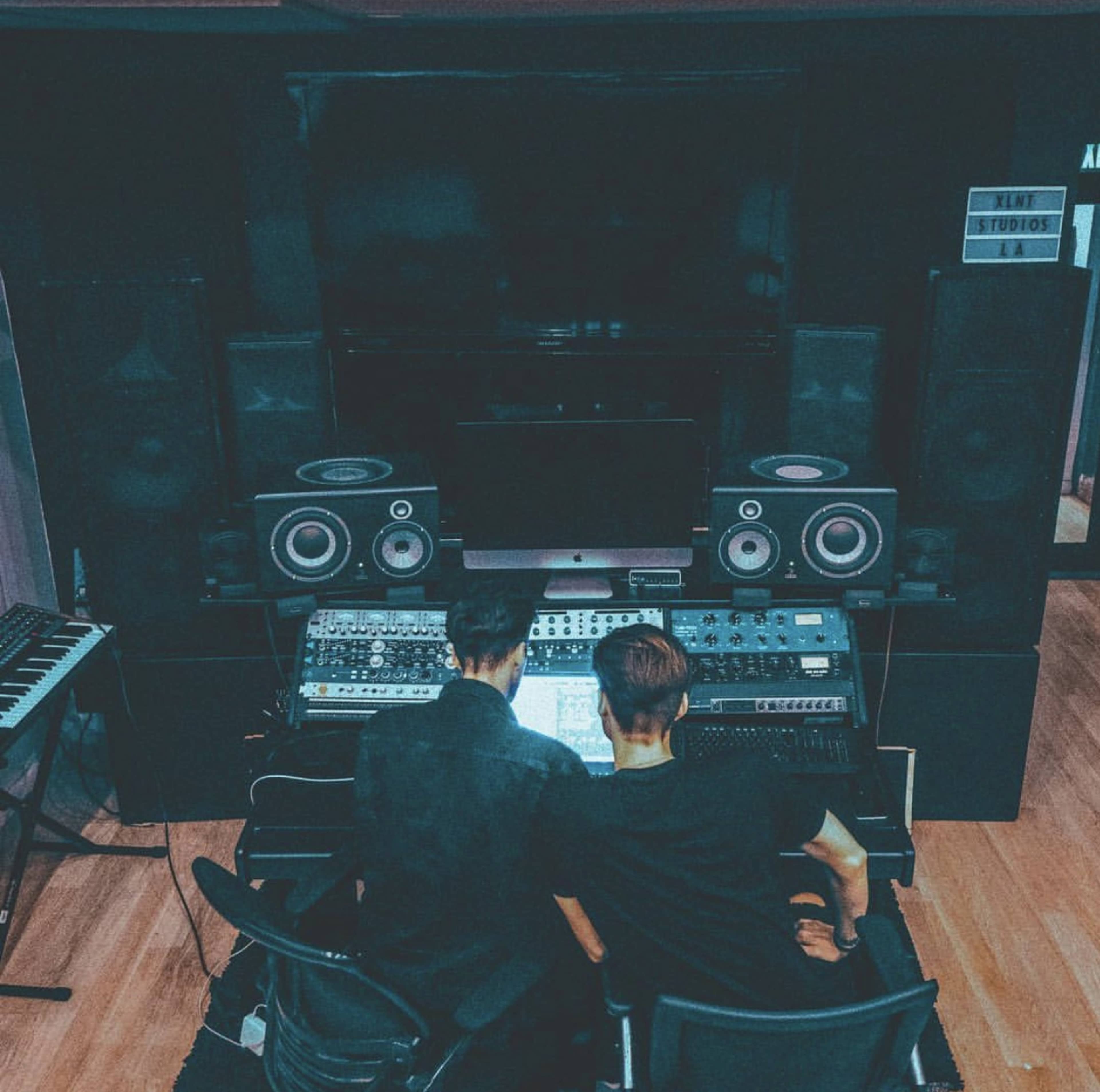 Two individuals are seated at a mixing desk in a music studio, surrounded by audio equipment and speakers.