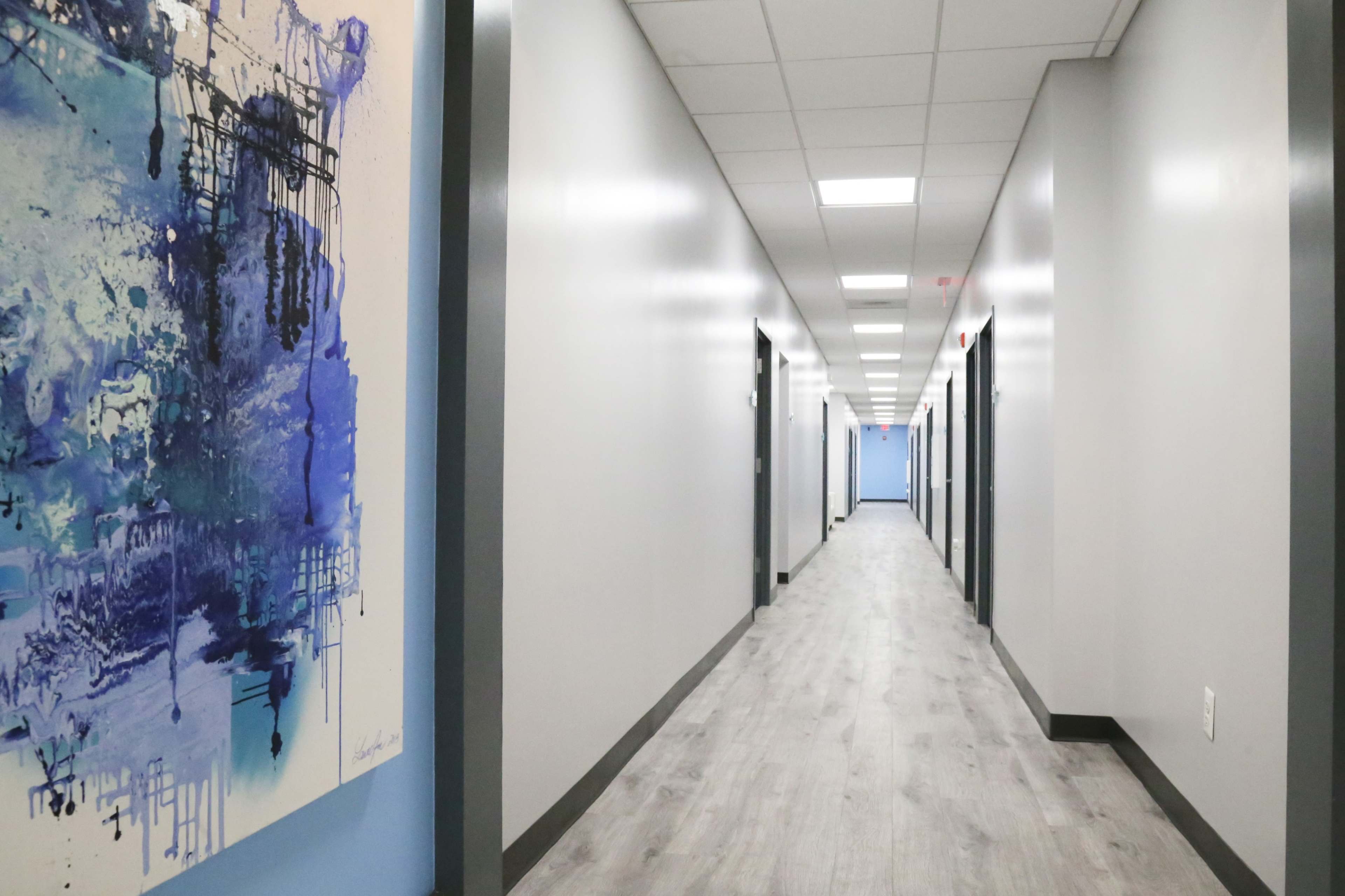 A long, well-lit hallway with light gray walls, a wooden floor, and doors on either side, along with a large abstract painting in blue and white at the entrance.