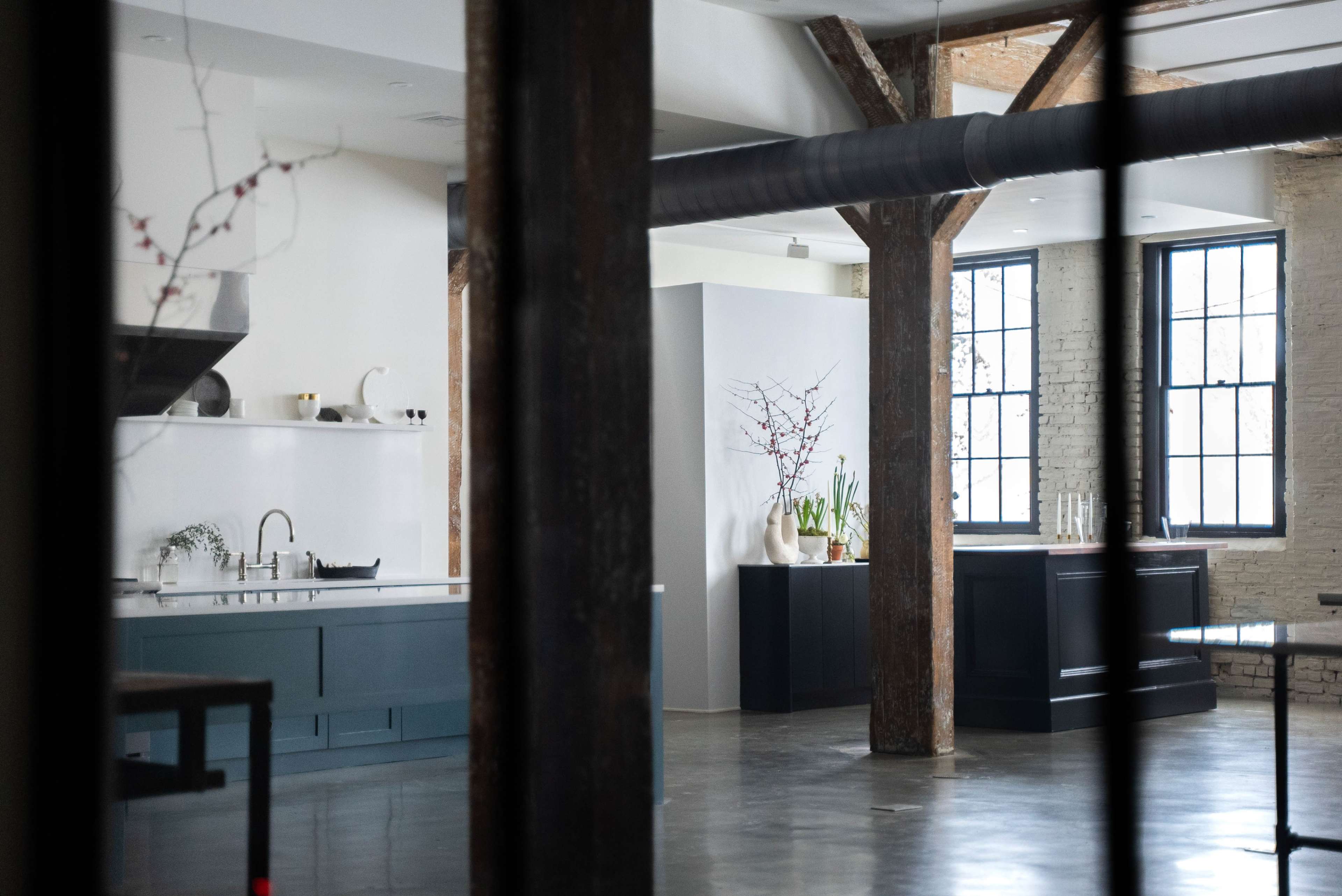 A modern kitchen with wooden beams, a large island, and shelves displaying ceramics and plants.