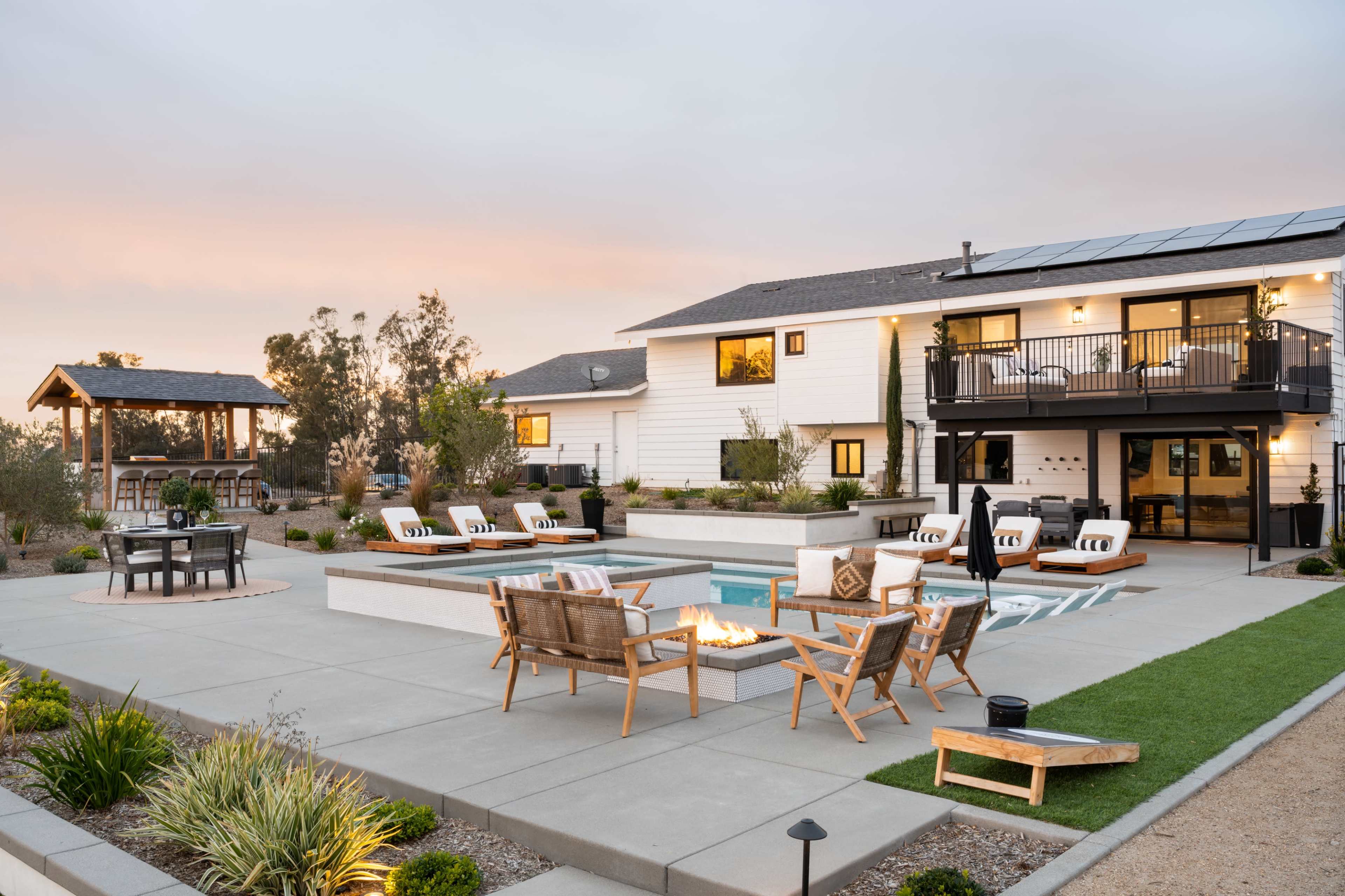 A modern backyard with a swimming pool, fire pit, and outdoor seating arranged around a concrete patio next to a two-story house.