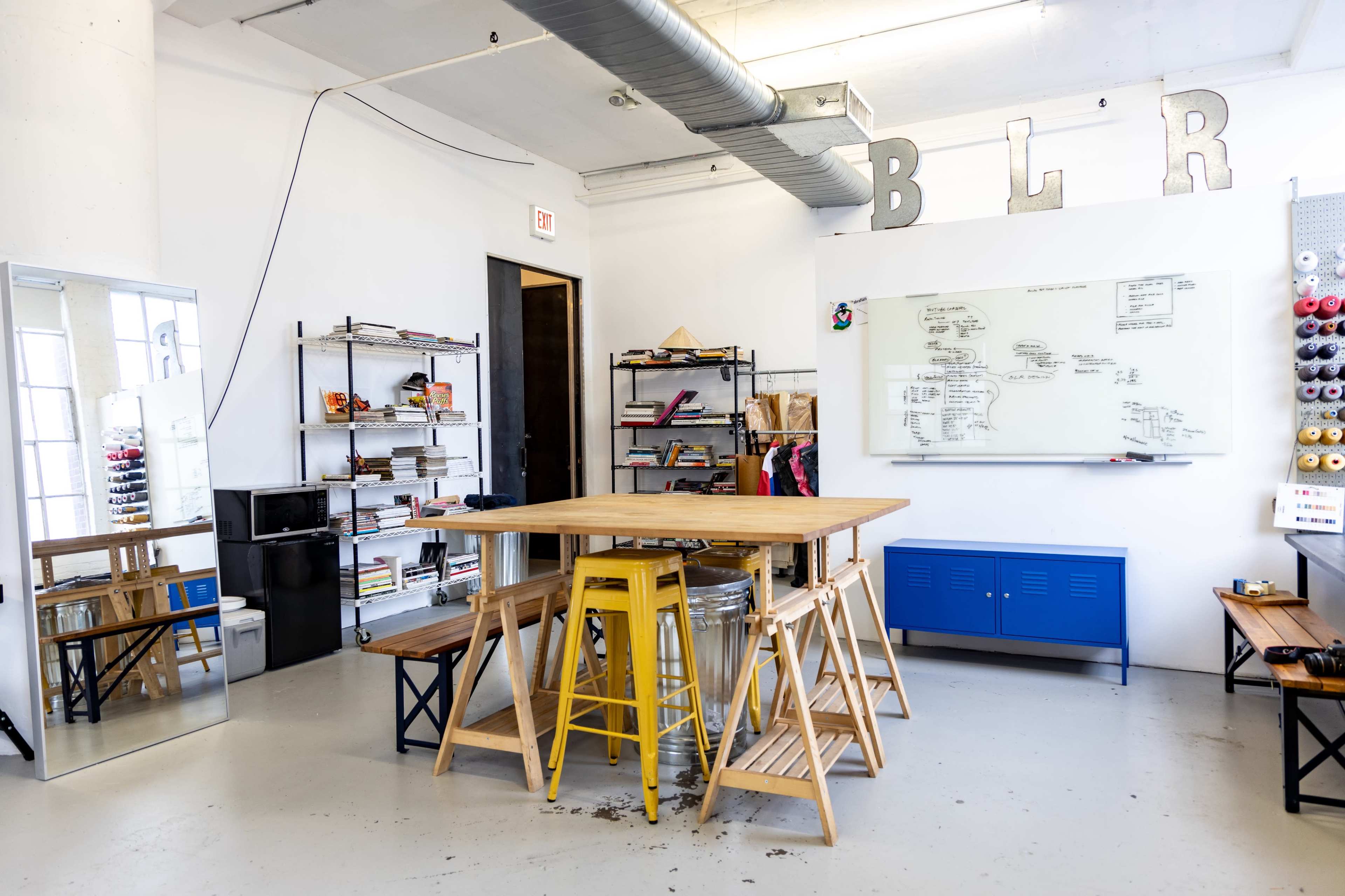 The room features a large wooden table surrounded by stools, a whiteboard on one wall, shelves with books and materials, and a mirror reflecting the space.