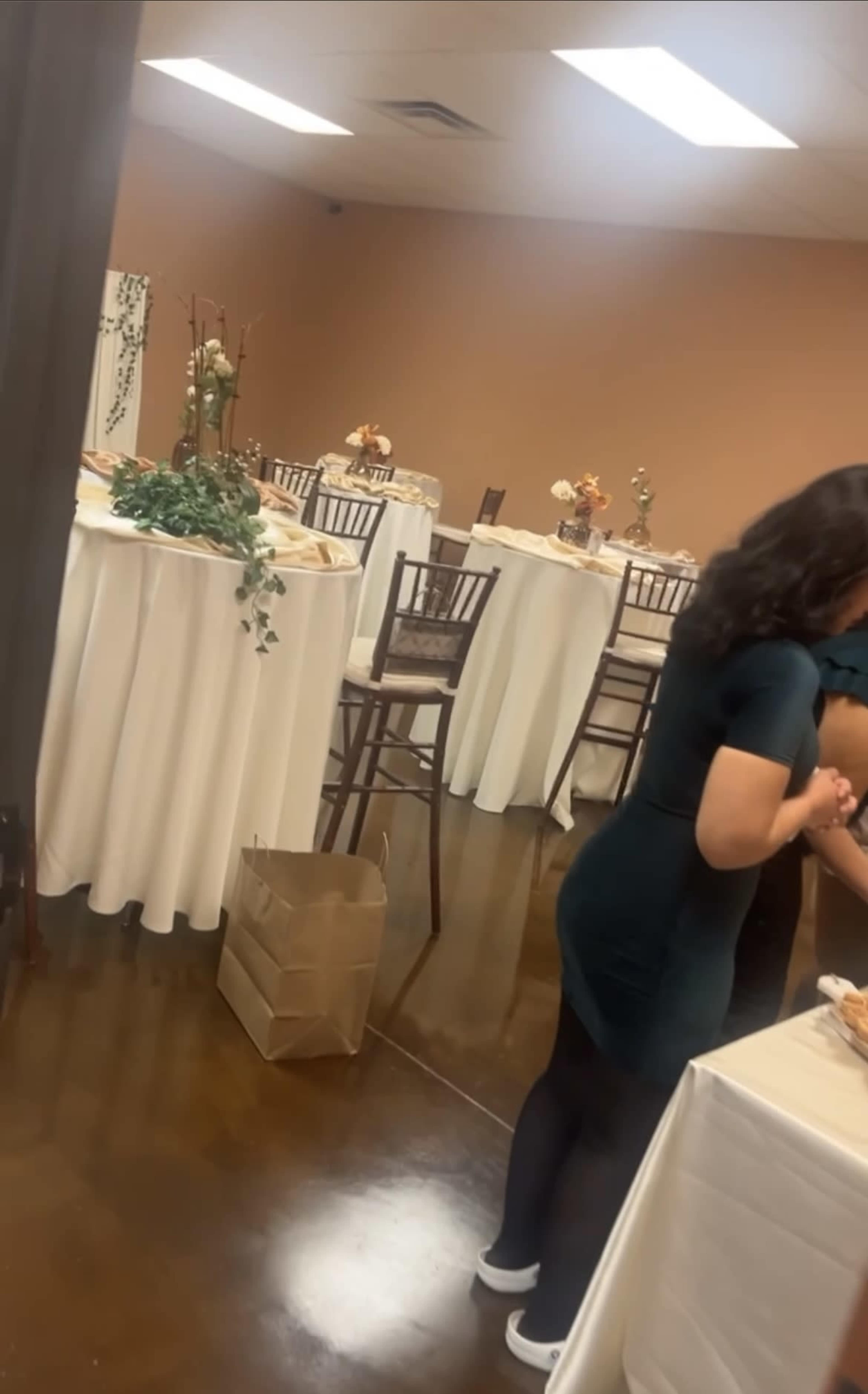 The image shows a decorated event space with tables covered in white tablecloths and a woman in a green dress standing near a food display.