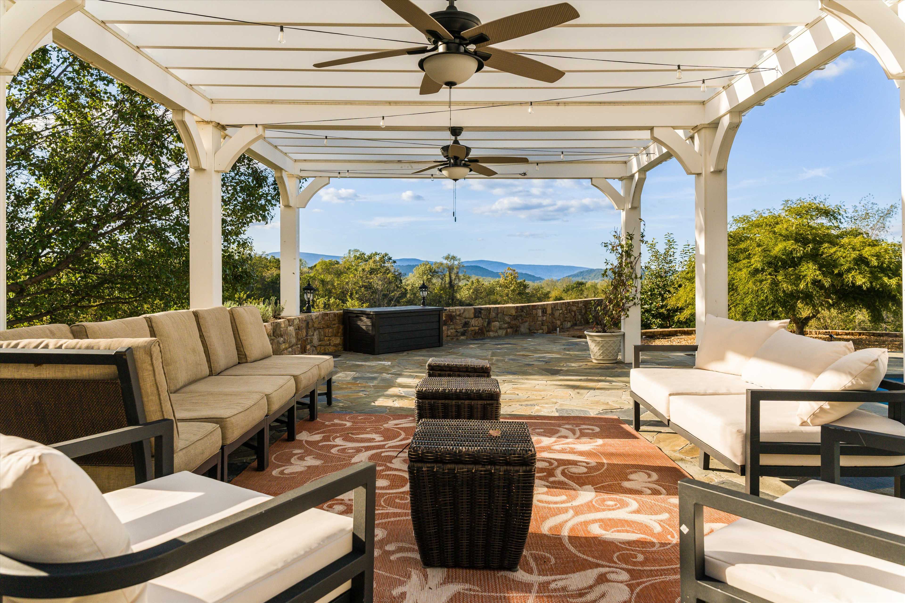 The image features a covered outdoor seating area with a stone floor and mountain views, furnished with beige couches and a central coffee table.