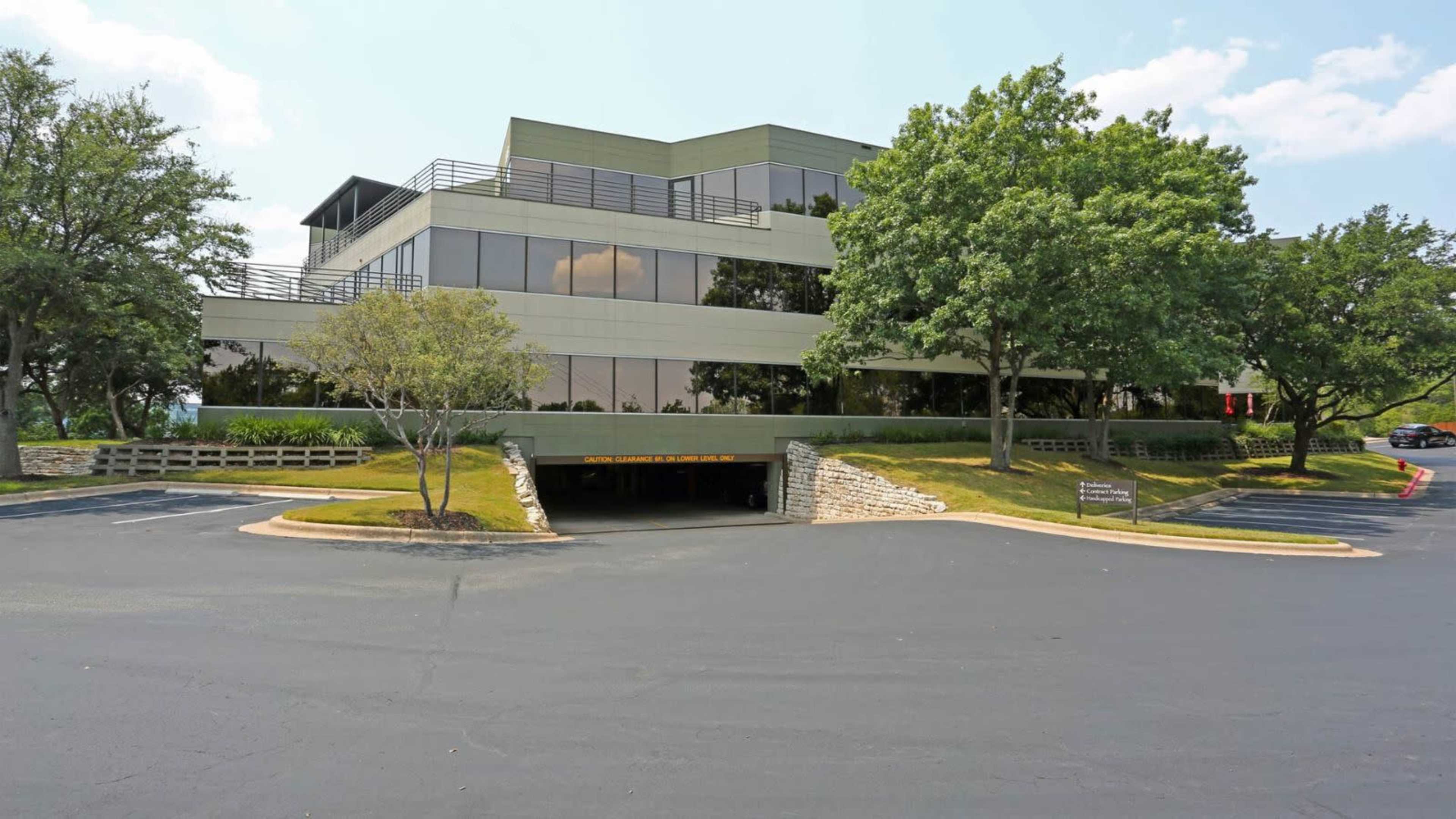A modern office building with large windows sits next to a parking lot and landscaped trees.