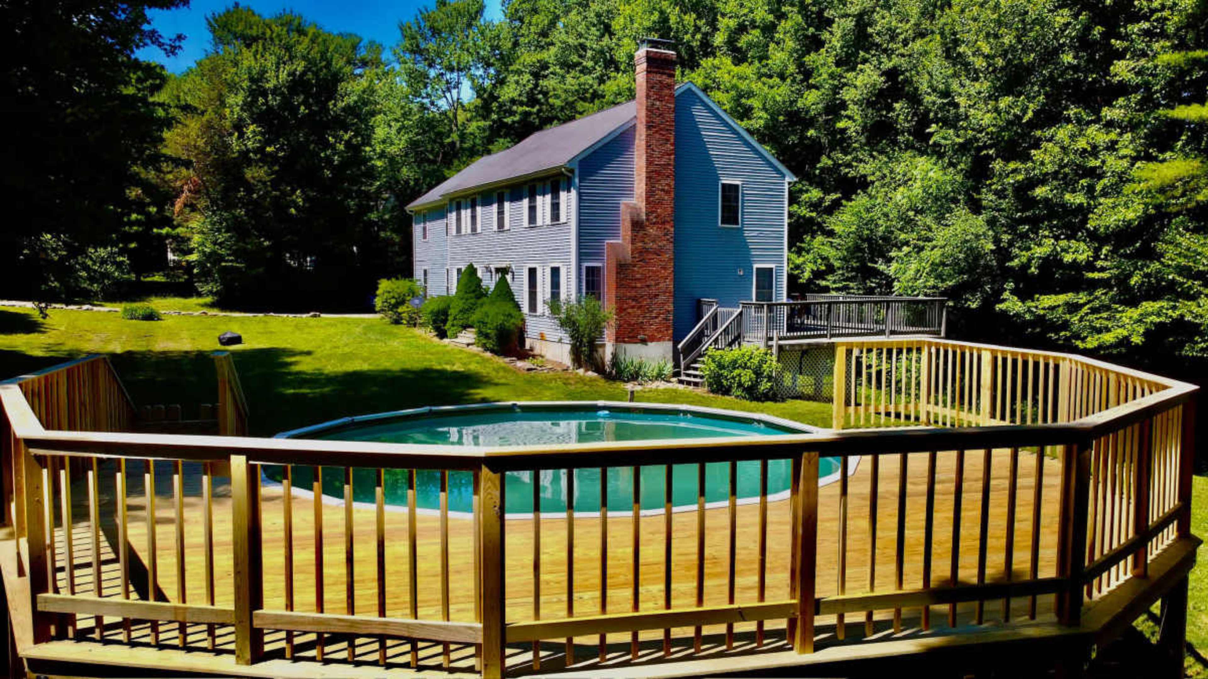 A two-story blue house is situated near a circular above-ground pool surrounded by a wooden deck in a green, wooded area.