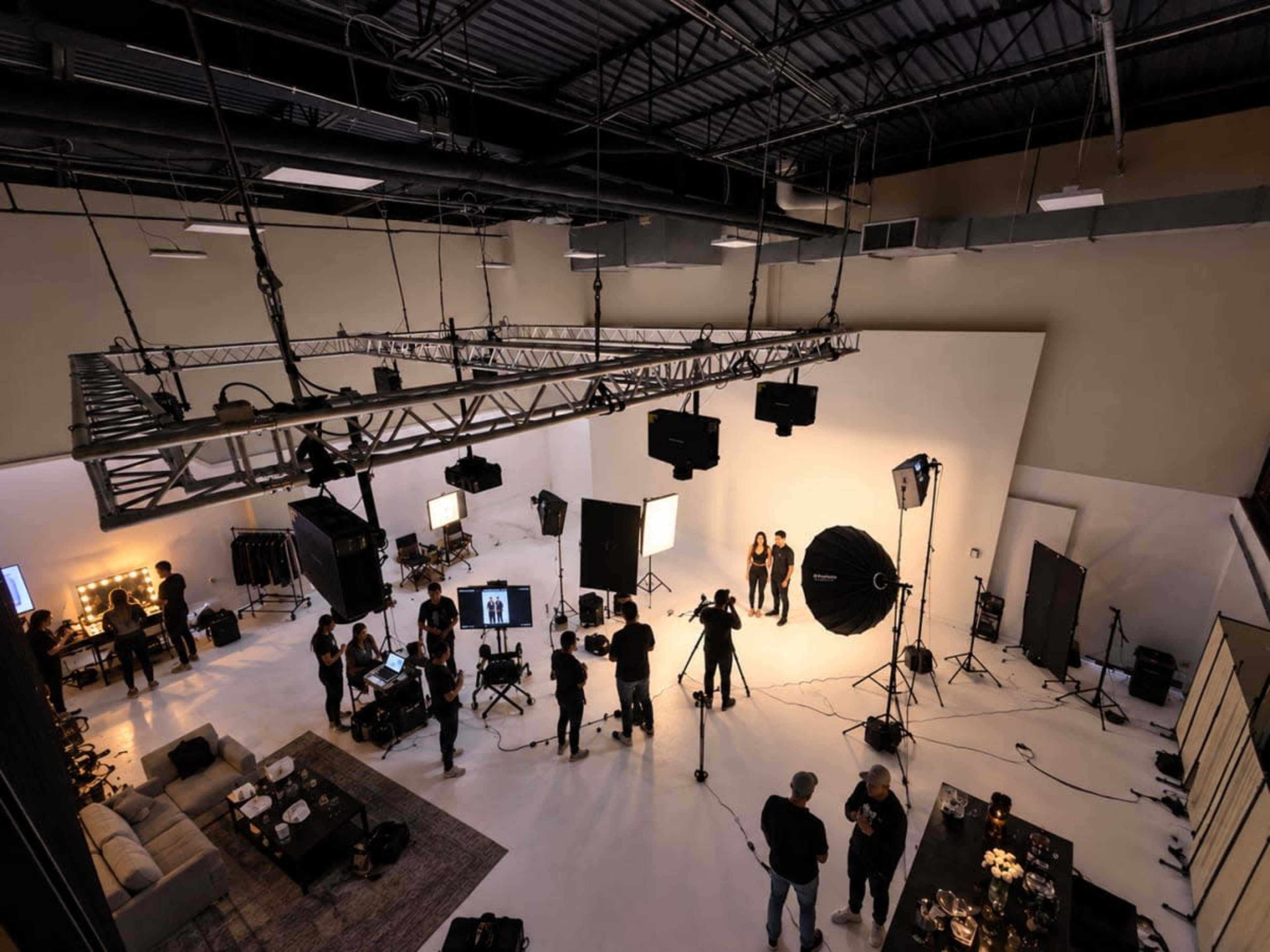 A large photography studio is set up with multiple lights and equipment as crew members work around a backdrop where two models are posing.