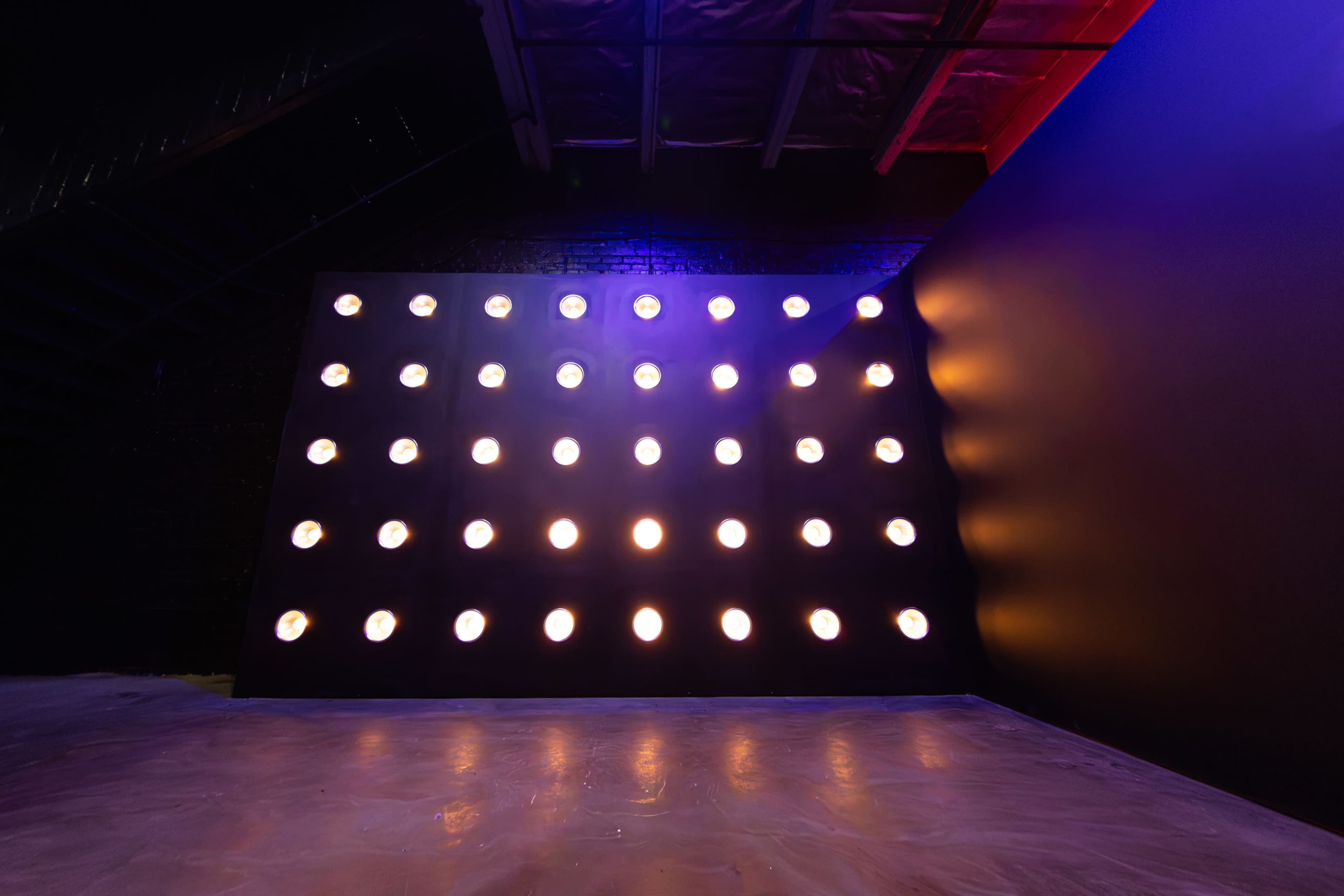 A wall with rows of brightly lit circular fixtures is positioned against a dark background in an empty room.