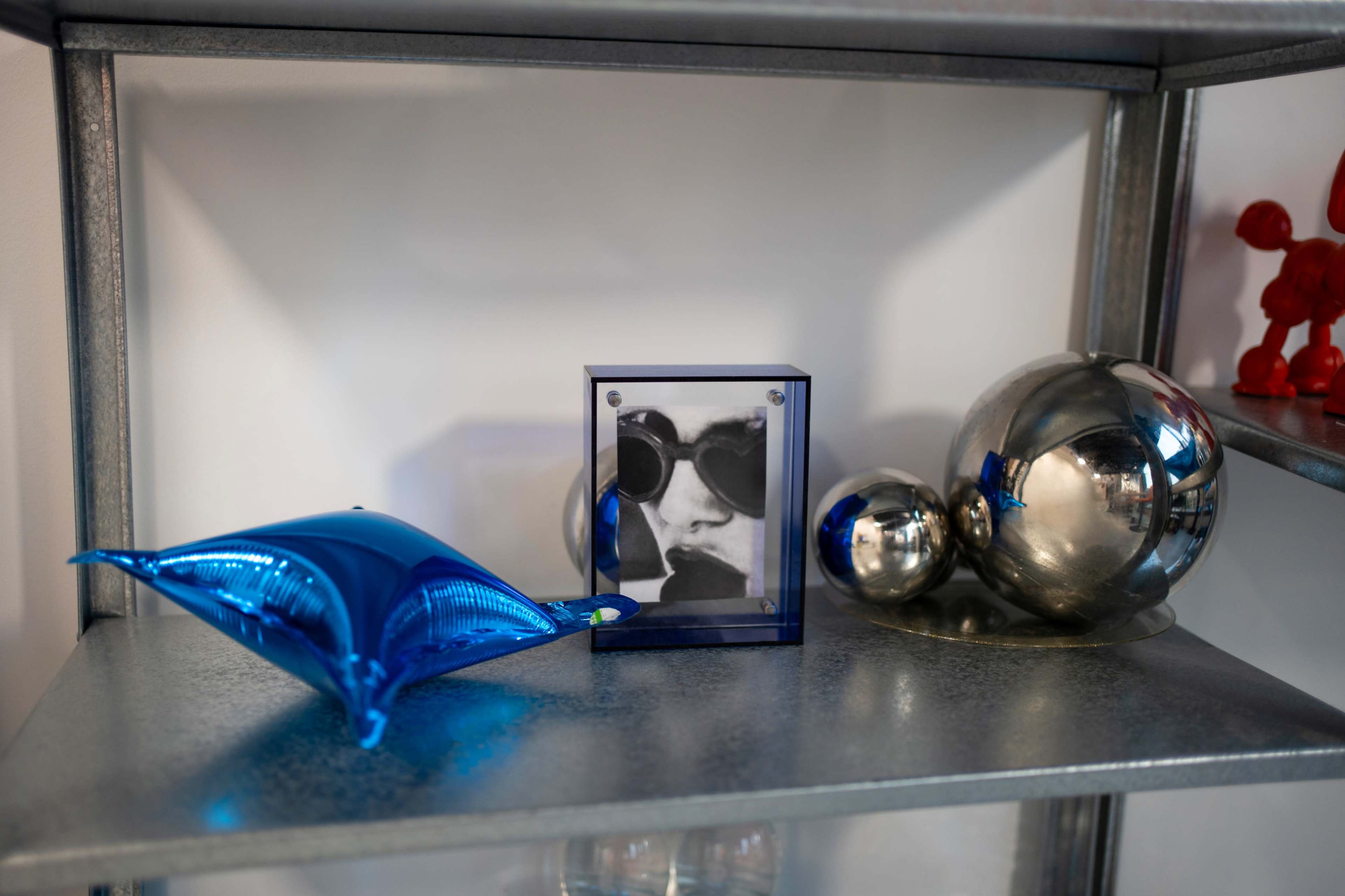 The image shows a metal shelf displaying a blue decorative pillow, a framed black and white photo of a person wearing sunglasses, and several shiny silver spheres.