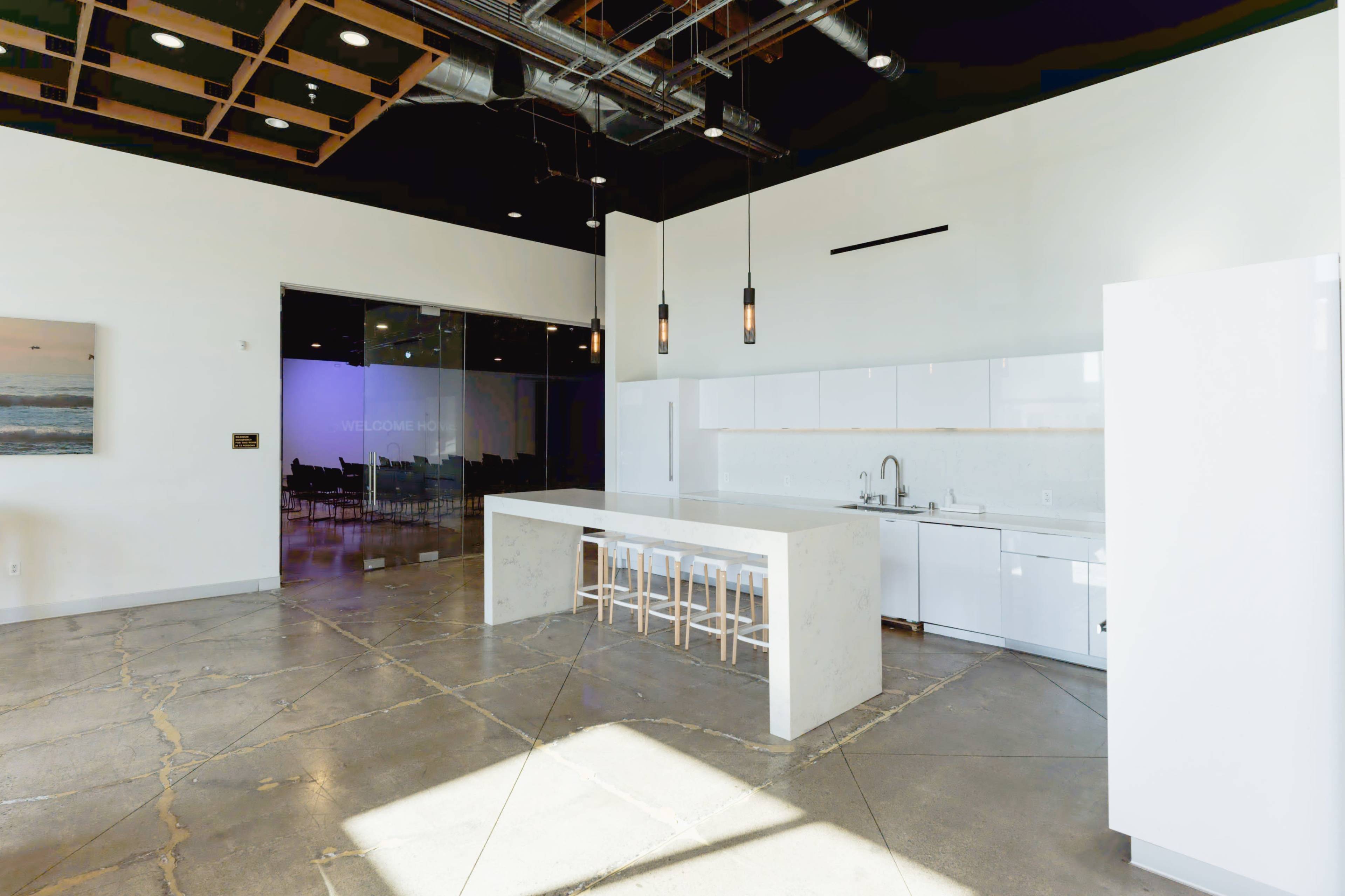 The image shows a modern, spacious kitchen area featuring a large island with bar stools, adjacent to a sleek white cabinetry setup, and large windows that offer a view of a well-lit event space.