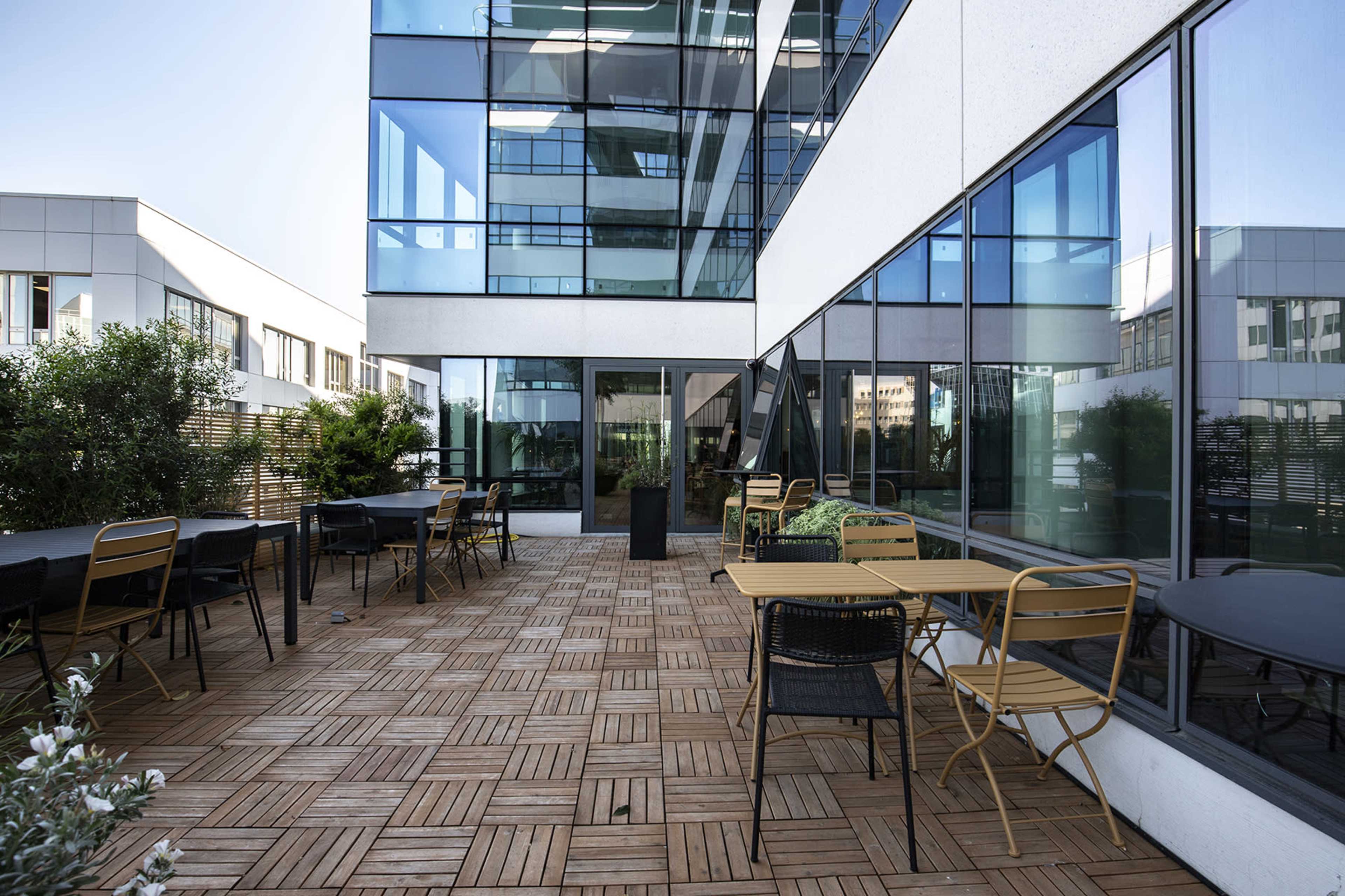 An outdoor terrace area with wooden flooring, surrounded by glass walls and equipped with several tables and chairs.