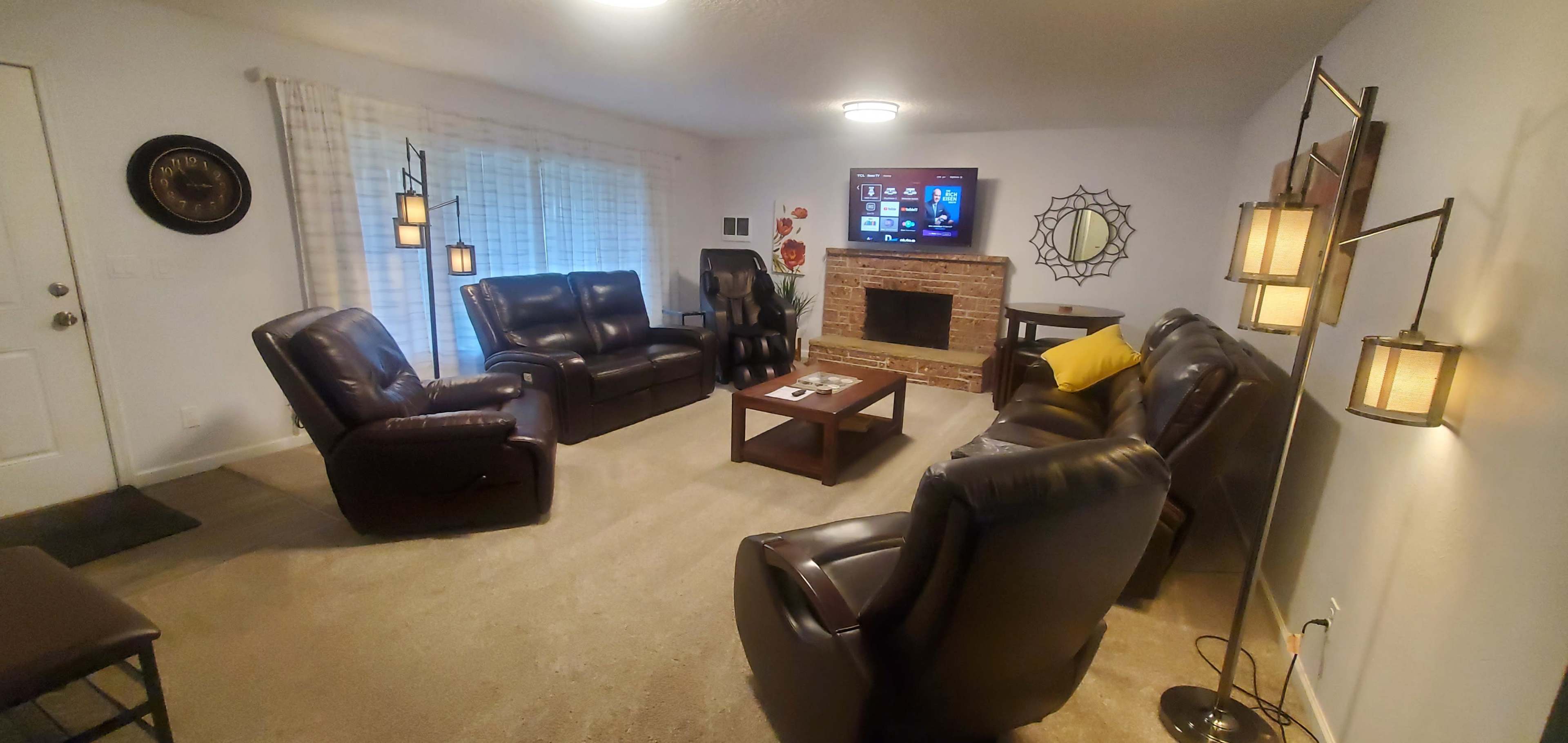A living room features two recliners, a sofa, a coffee table, and a television mounted above a brick fireplace, with a large window covered by light curtains.