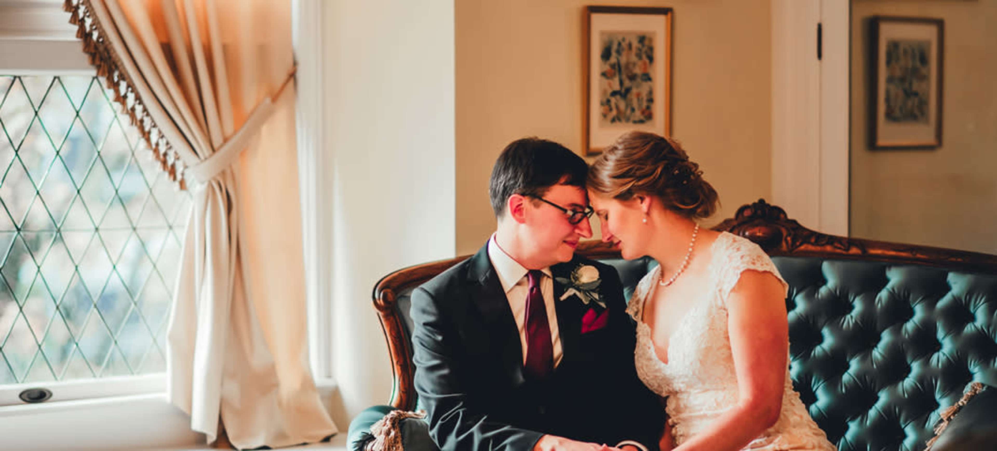 A couple sits closely together on a vintage couch, sharing an intimate moment in a well-lit room.