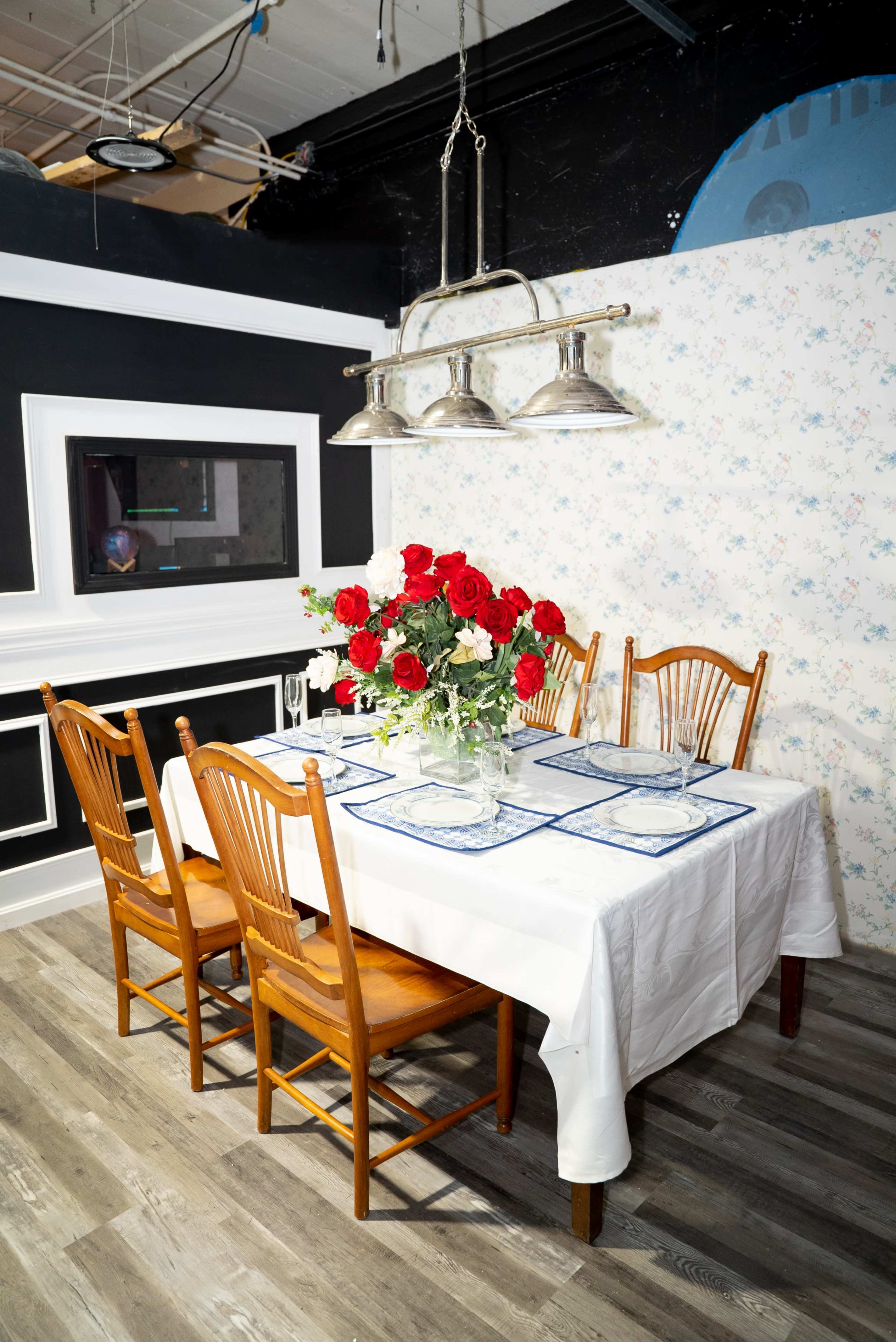 A dining table set with a white tablecloth, blue placemats, and a vase of red roses is surrounded by wooden chairs in a room with patterned wallpaper.