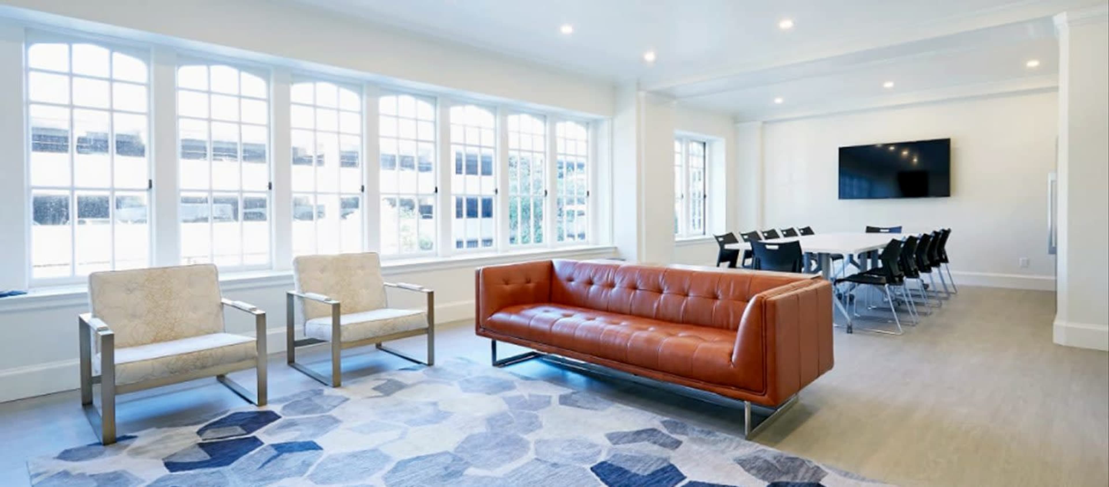 A modern office space featuring a brown leather sofa, two armchairs, a large rug with a geometric pattern, and a conference table surrounded by black chairs, all in a well-lit room with multiple windows.