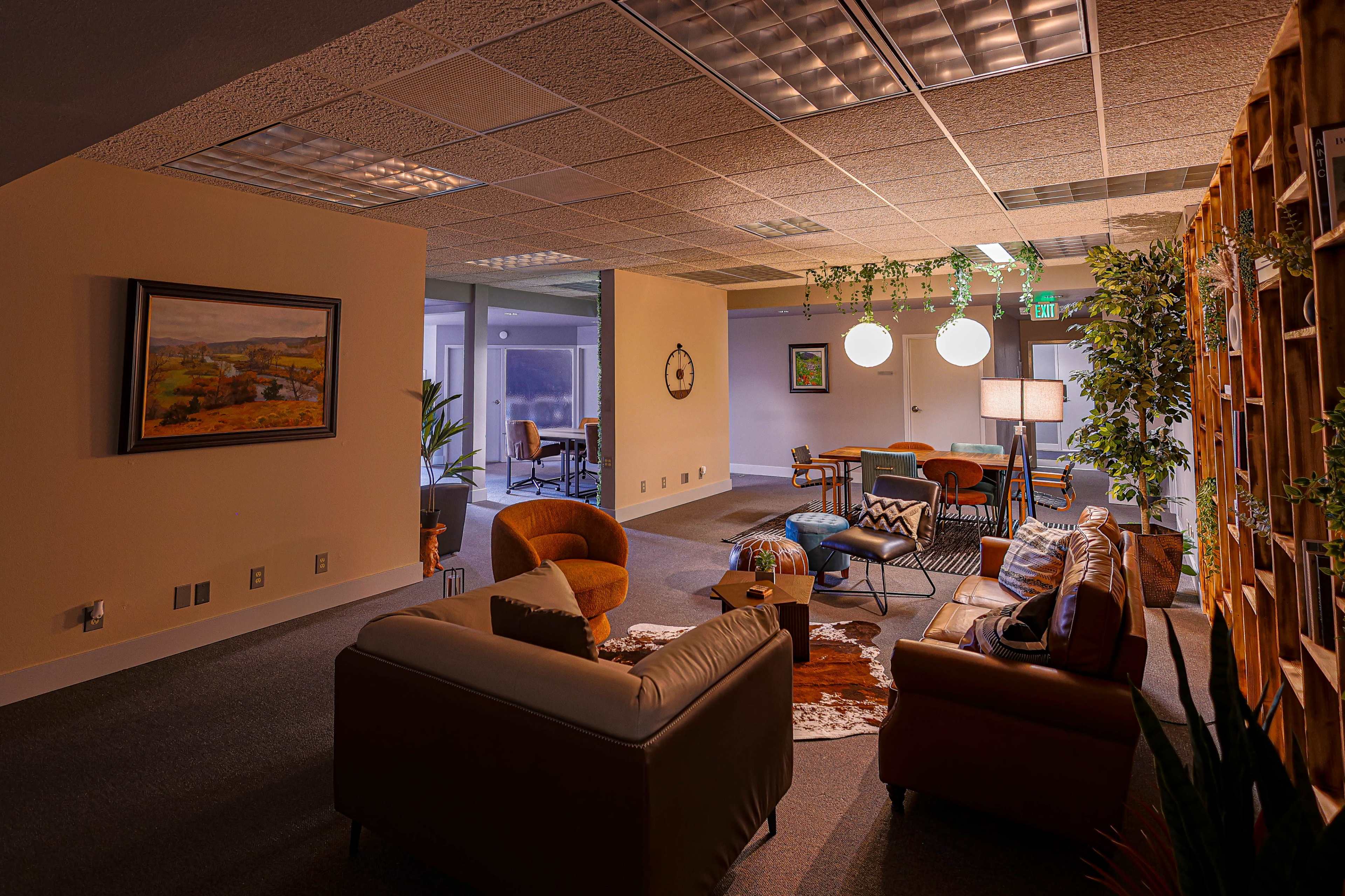 The image shows a modern office lounge area with comfortable seating, a bookshelf, decorative plants, and overhead lighting.